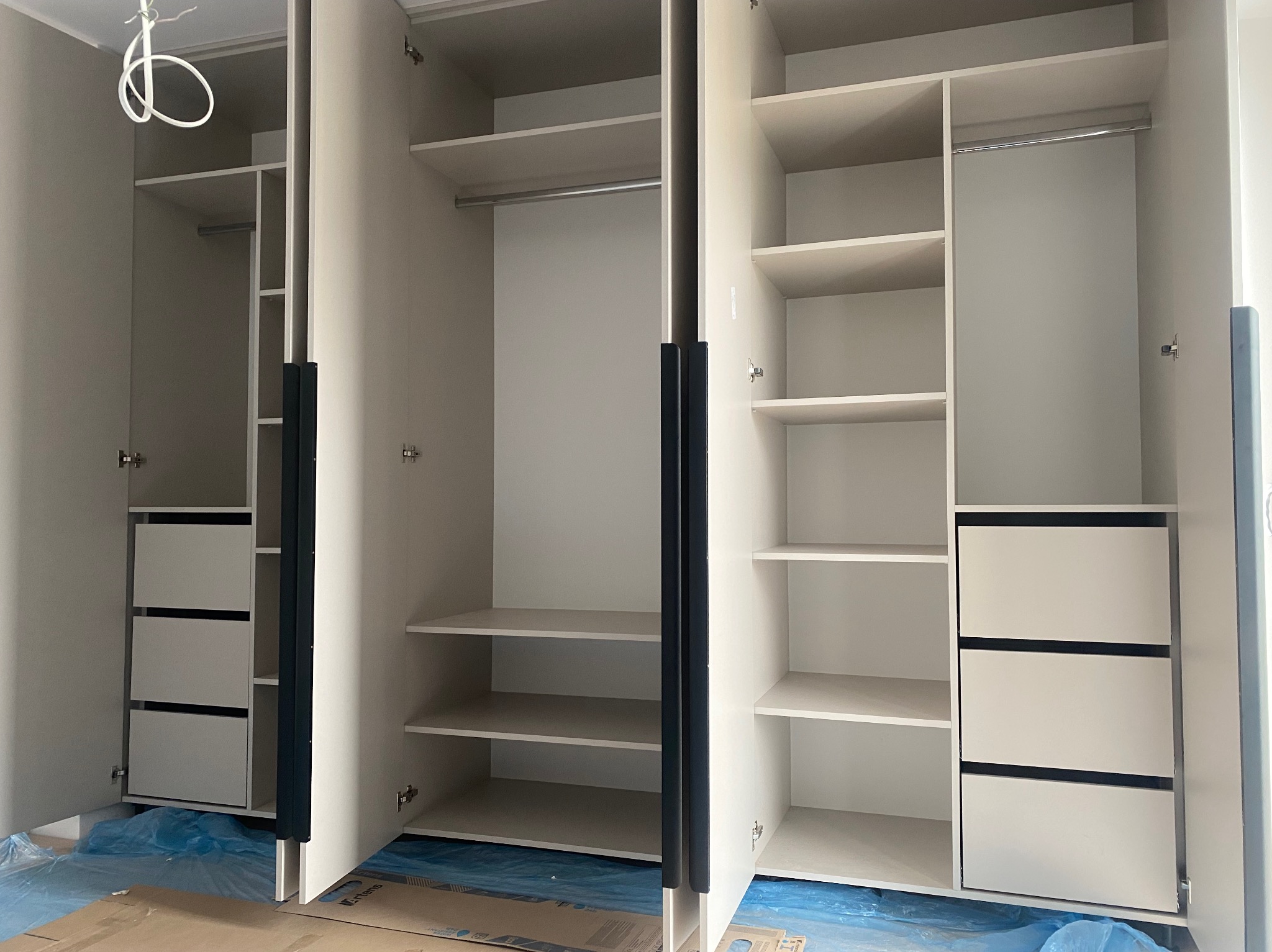 Jasna szafa na wymiar z szufladami i półkami, widok wnętrza, uchwyty w kolorze grafitowym. Podłoga zabezpieczona folią malarską, co sugeruje świeży montaż.