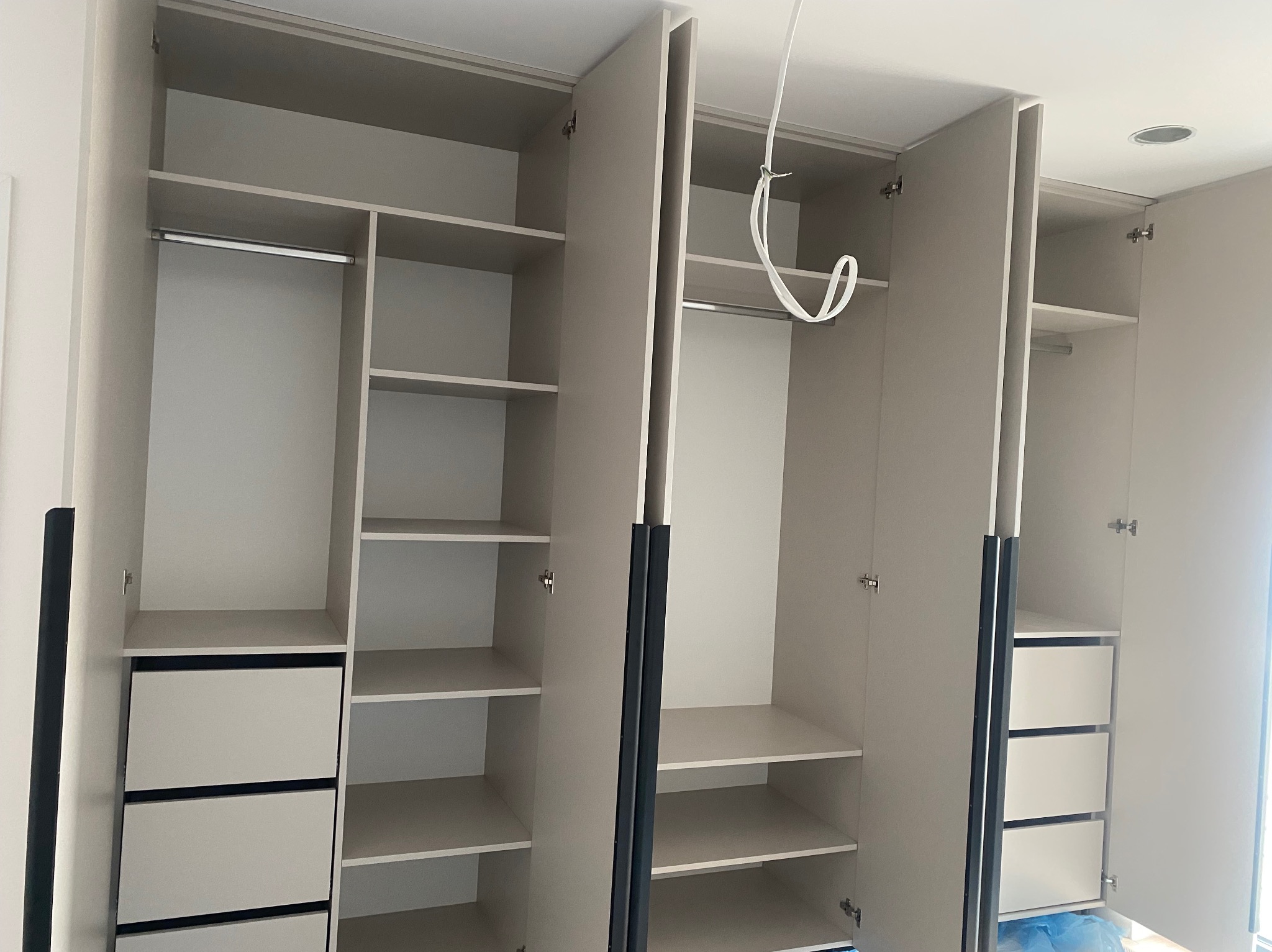 Jasna szafa na wymiar z szufladami i półkami, otwarte drzwi ukazują wnętrze. Widoczne okucia i elementy montażowe. Minimalistyczny design, oświetlenie sufitowe.