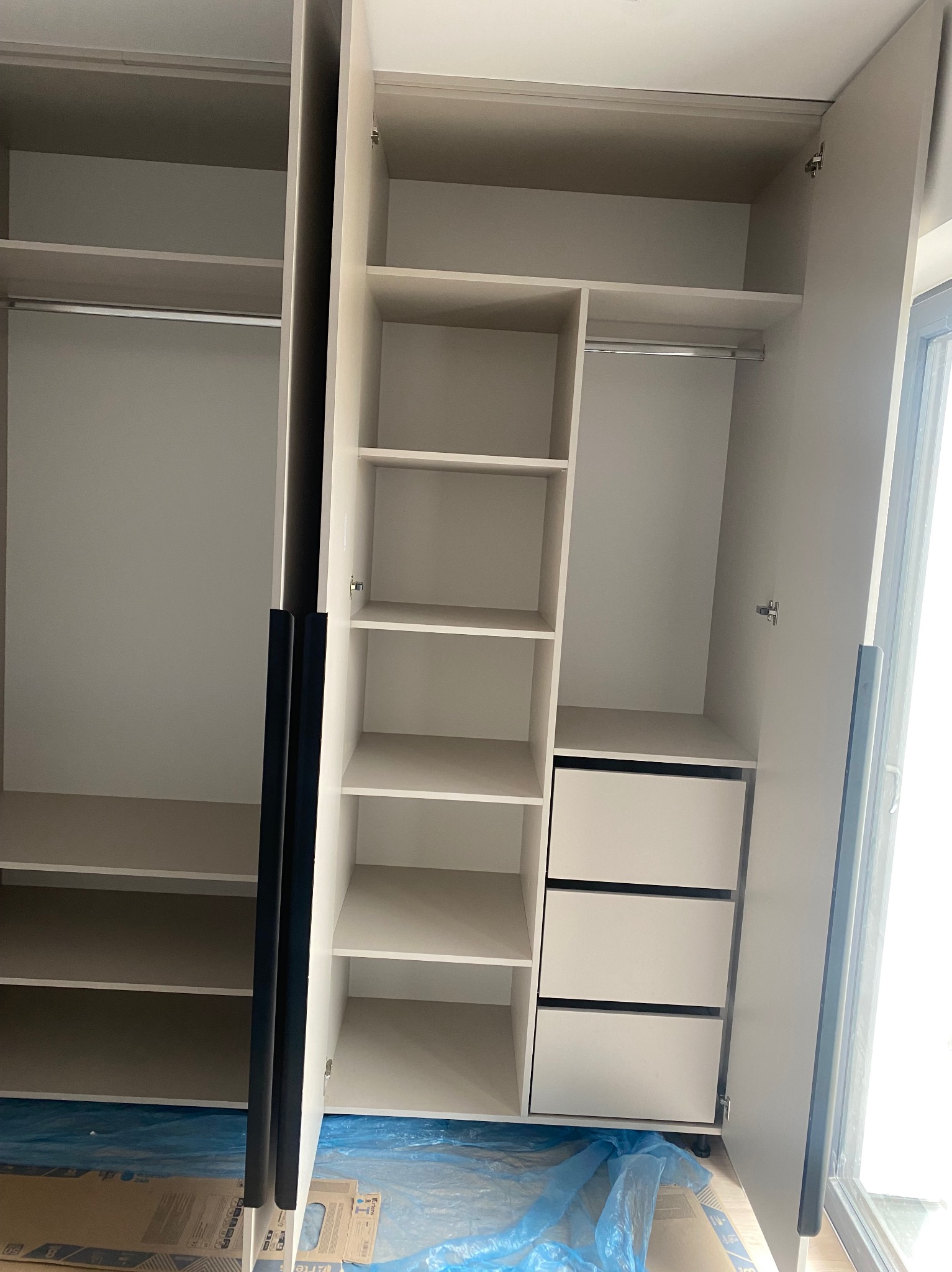 Jasna szafa na wymiar z otwartymi drzwiami, widoczne półki i szuflady. Minimalistyczny design, czarne uchwyty. Podłoga zabezpieczona folią ochronną.