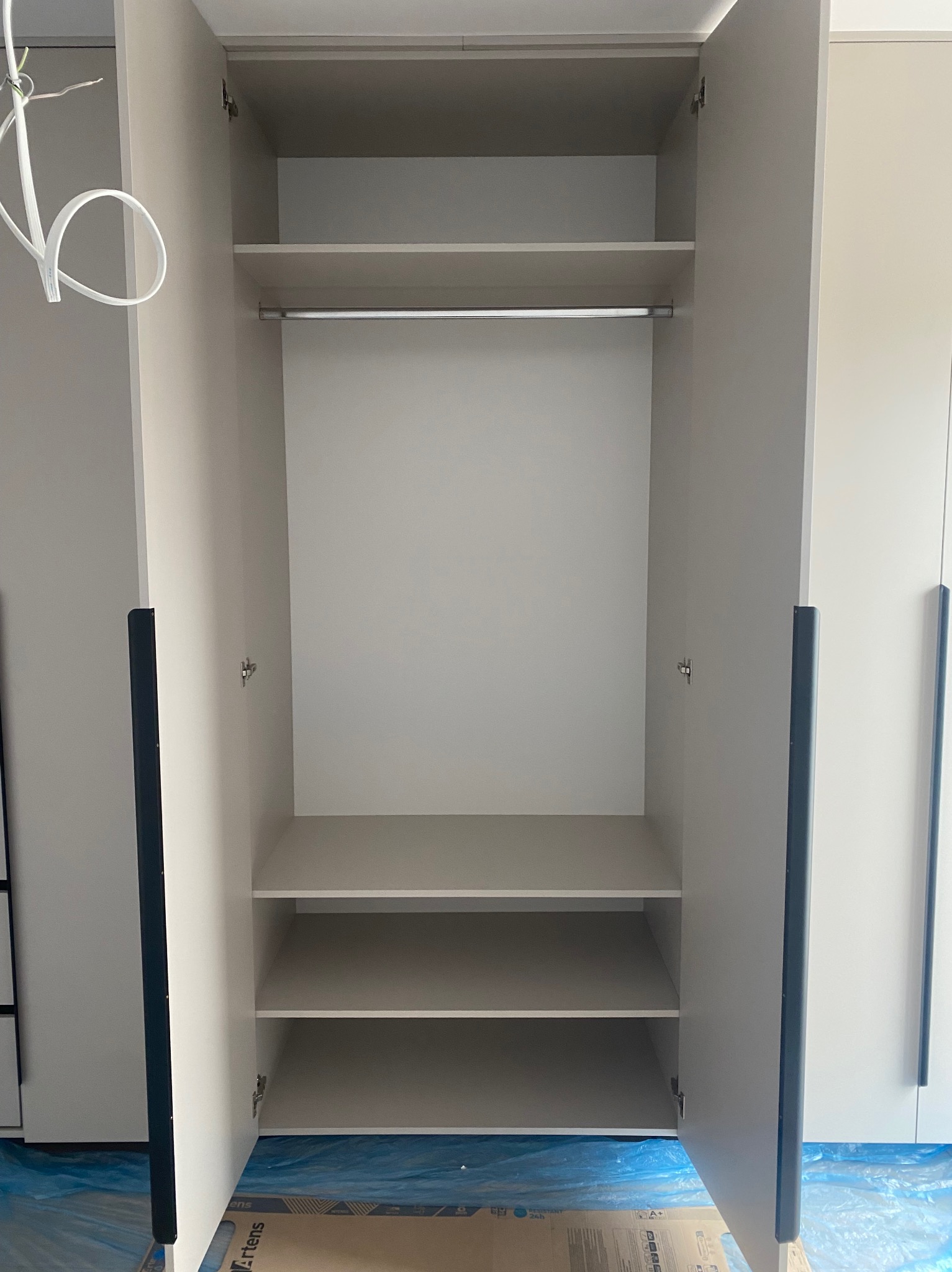 Wnętrze szafy z otwartymi drzwiami, widoczne półki i drążek na ubrania. Jasne kolory, minimalistyczny design. Na podłodze folia ochronna.