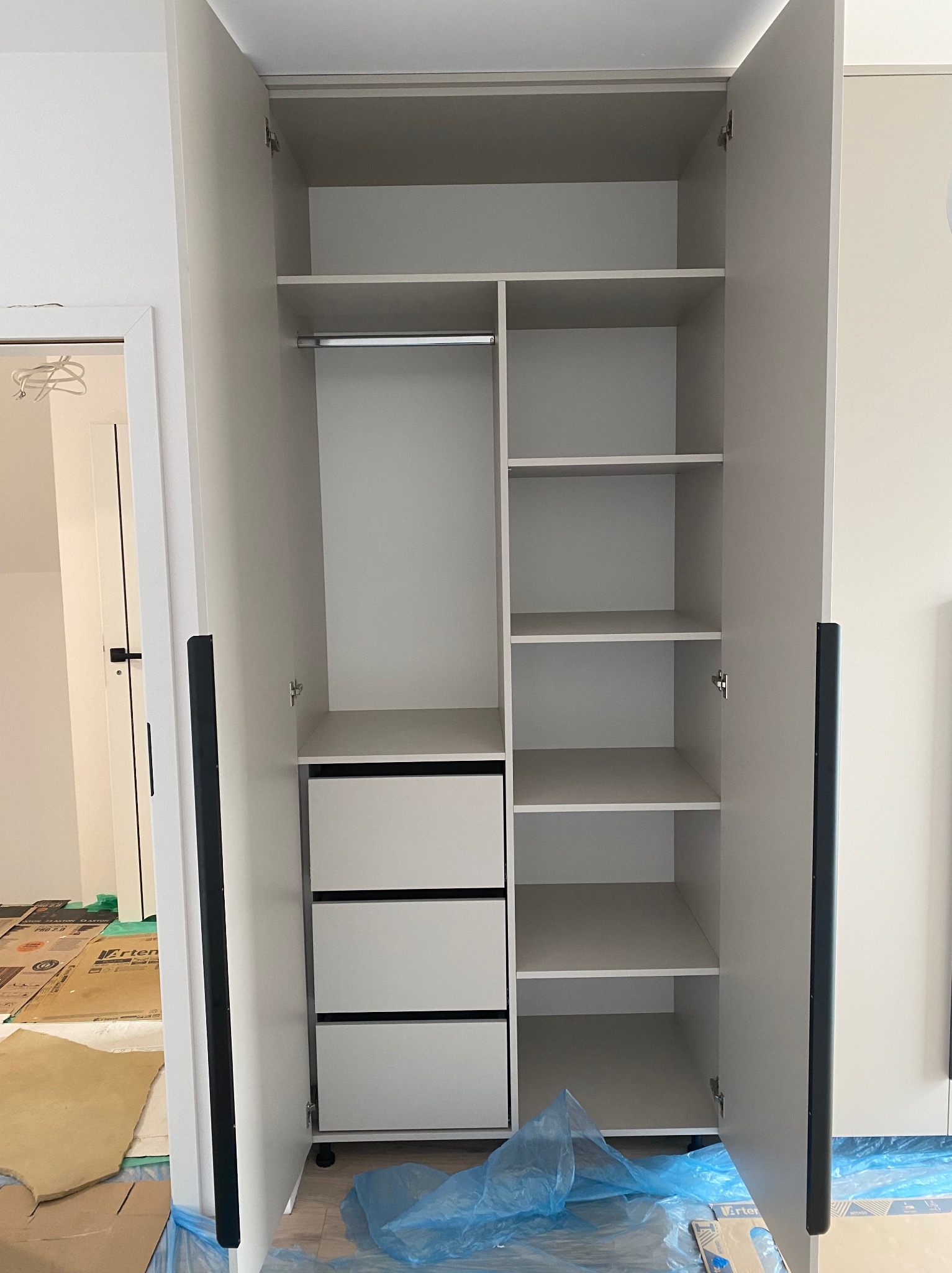 Jasna szafa z otwartymi drzwiami, z półkami i szufladami. Widoczne zabezpieczenia podłogi, co sugeruje montaż lub prace wykończeniowe. Minimalistyczny design, czarne uchwyty.