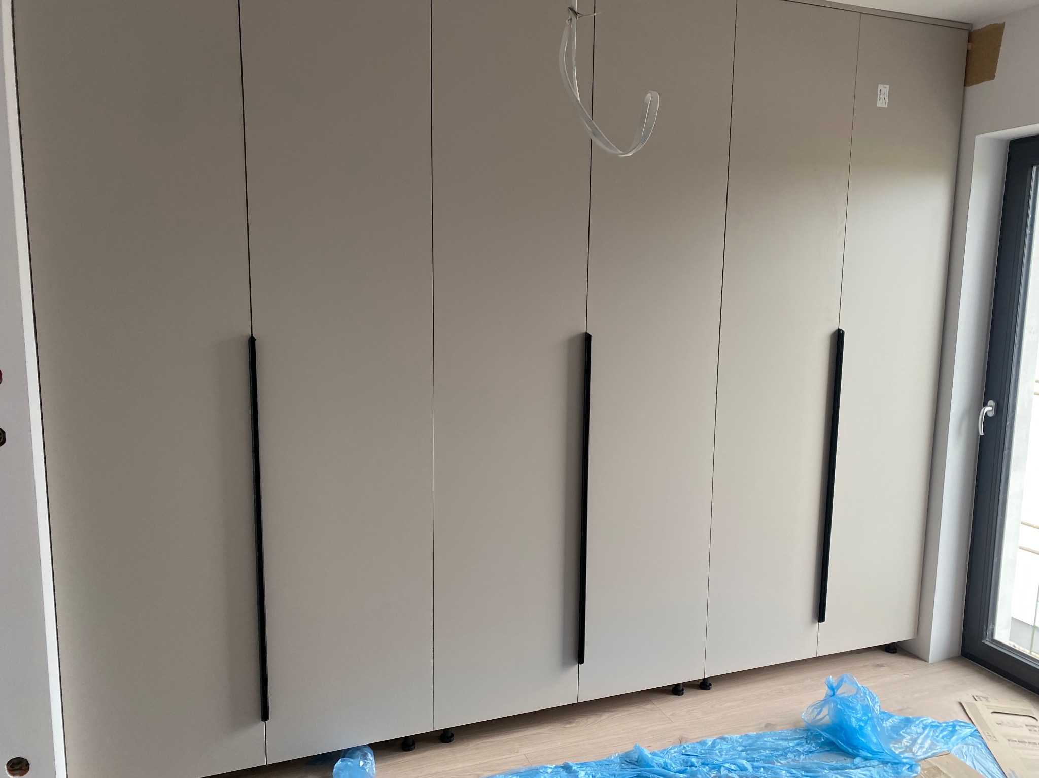 Jasna szafa na wymiar z długimi, czarnymi uchwytami, widoczna w nowym wnętrzu, częściowo zabezpieczona folią ochronną, z elementami instalacji na górze.