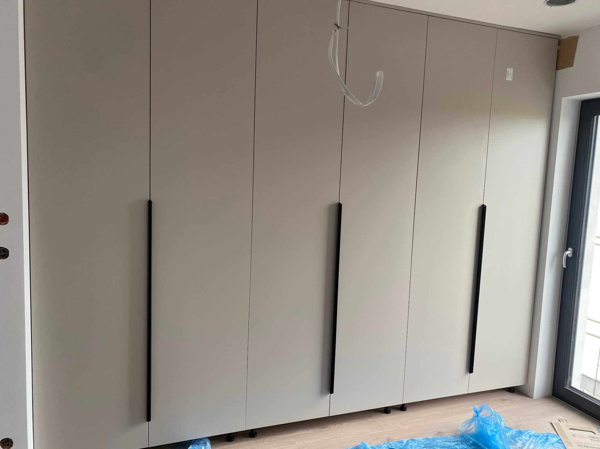 Duża szafa na wymiar z beżowymi frontami i długimi, czarnymi uchwytami, stojąca na drewnianej podłodze przy oknie. Widoczne zabezpieczenia podłogi folią.