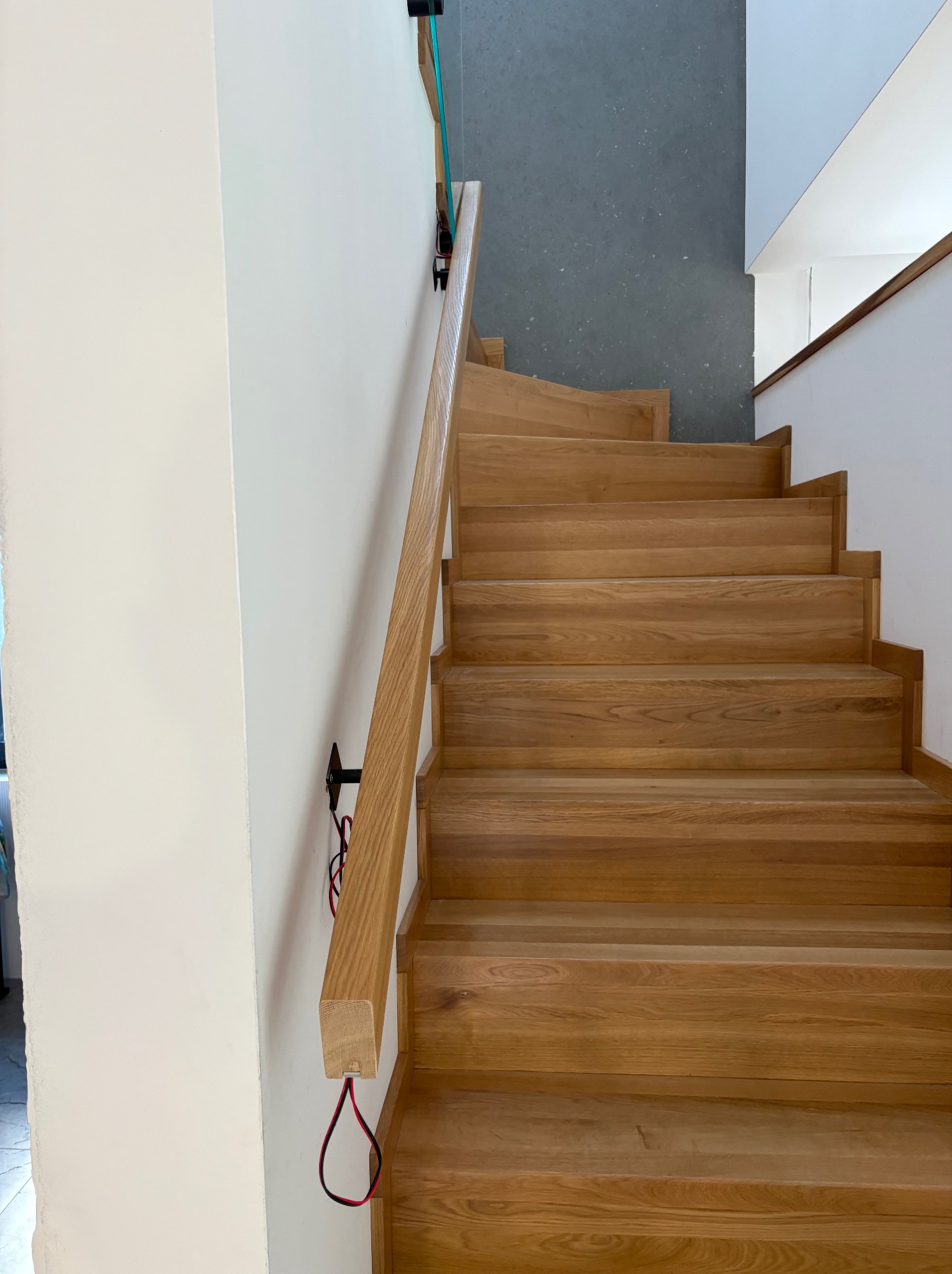 Jasne, drewniane schody z poręczą, zamontowaną na czarnych uchwytach z widocznymi przewodami, prowadzą w górę. Szare podłoże widoczne u góry. Minimalistyczny design.
