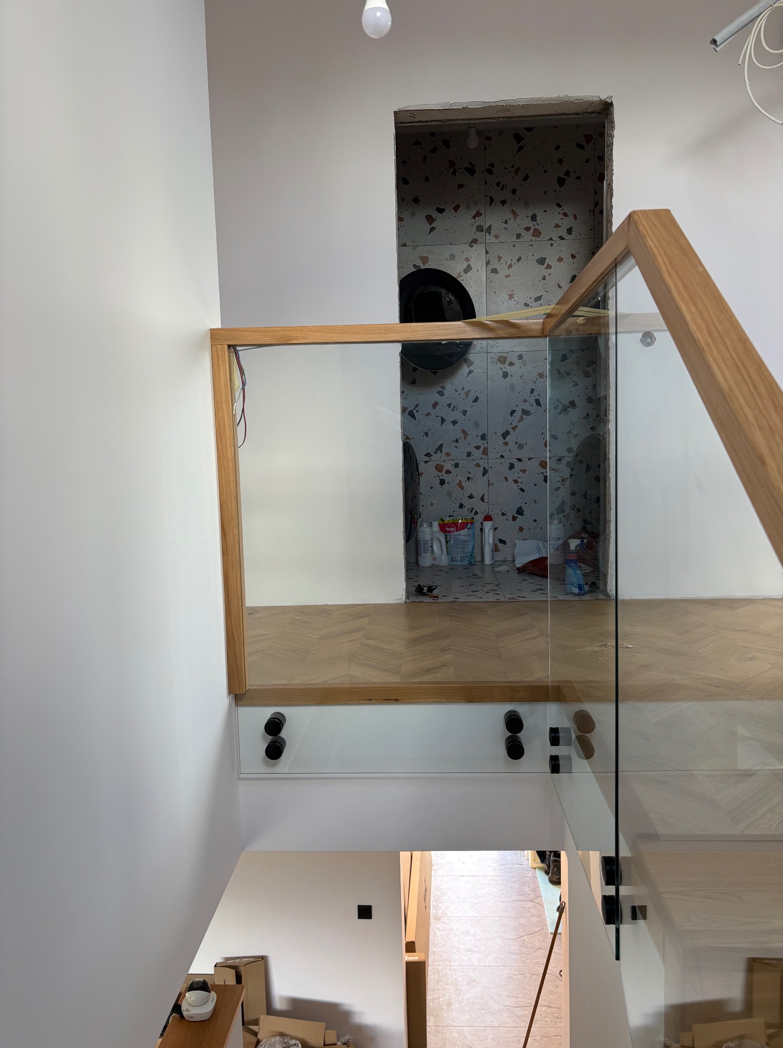 Szklana balustrada z drewnianymi elementami na schodach, widok z góry. Widoczne detale mocowania i wykończenia. W tle pomieszczenie w trakcie remontu.