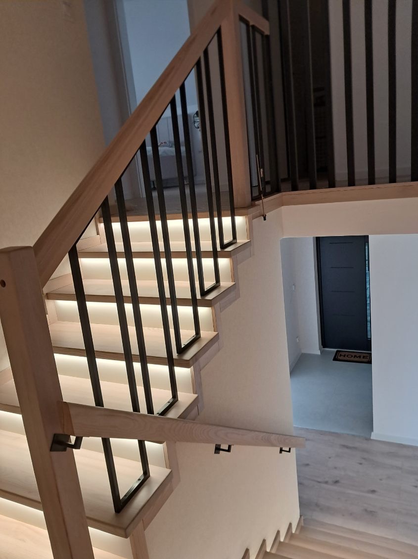 Nowoczesne schody drewniane z podświetleniem LED, czarnymi balustradami i jasnymi stopniami. Widok z góry na korytarz z czarnymi drzwiami.