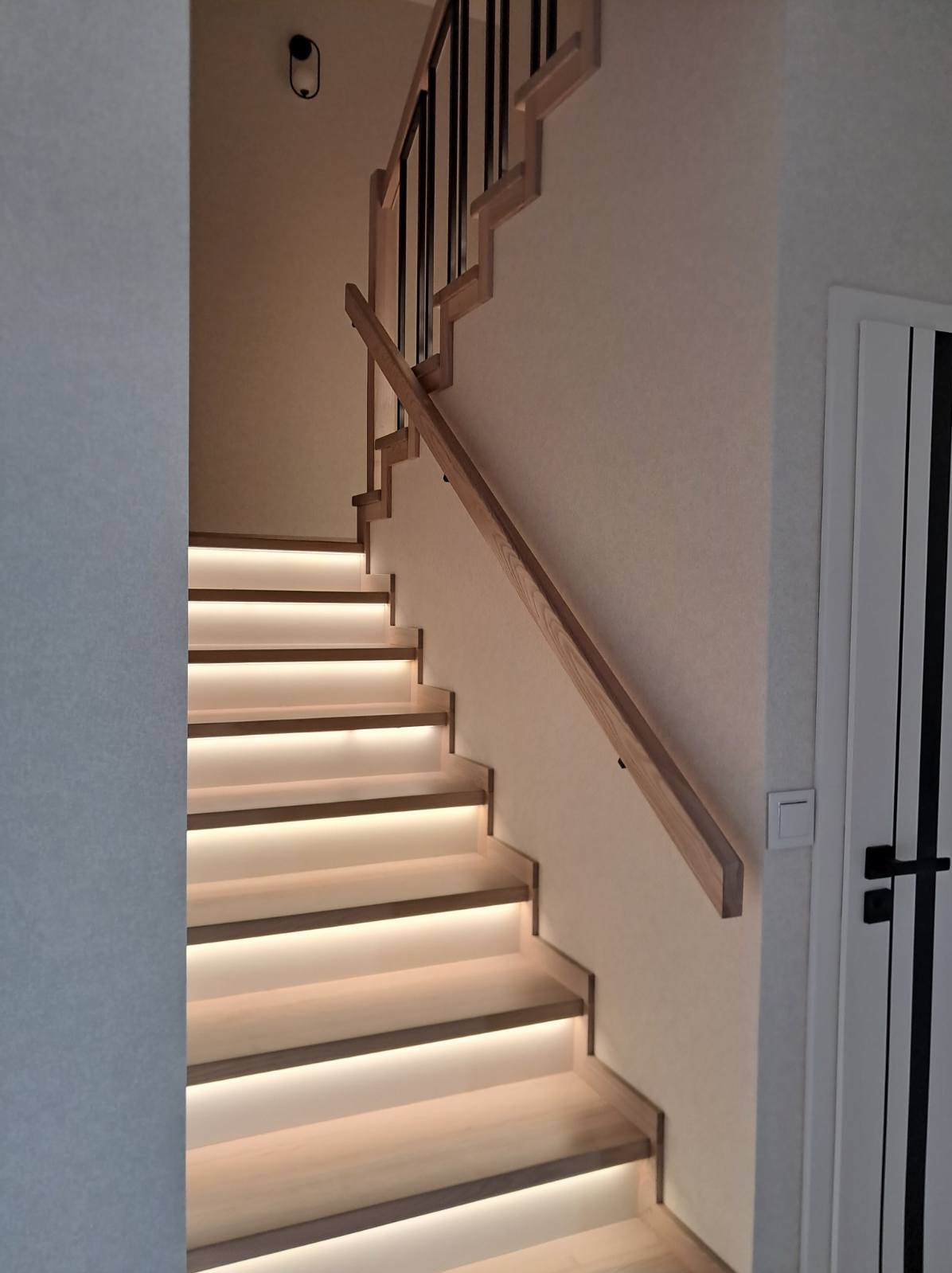 Klatka schodowa z drewnianymi schodami podświetlonymi ledami. Balustrada z drewnianą poręczą i czarnymi, metalowymi elementami. Białe ściany i drzwi z czarnymi akcentami.