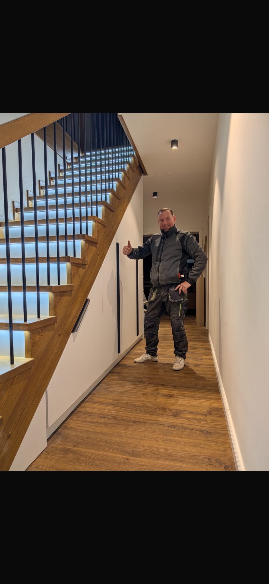 Nowoczesne schody drewniane z podświetleniem LED, czarnymi balustradami i uśmiechniętym stolarzem w tle, prezentujące efekt finalny realizacji.