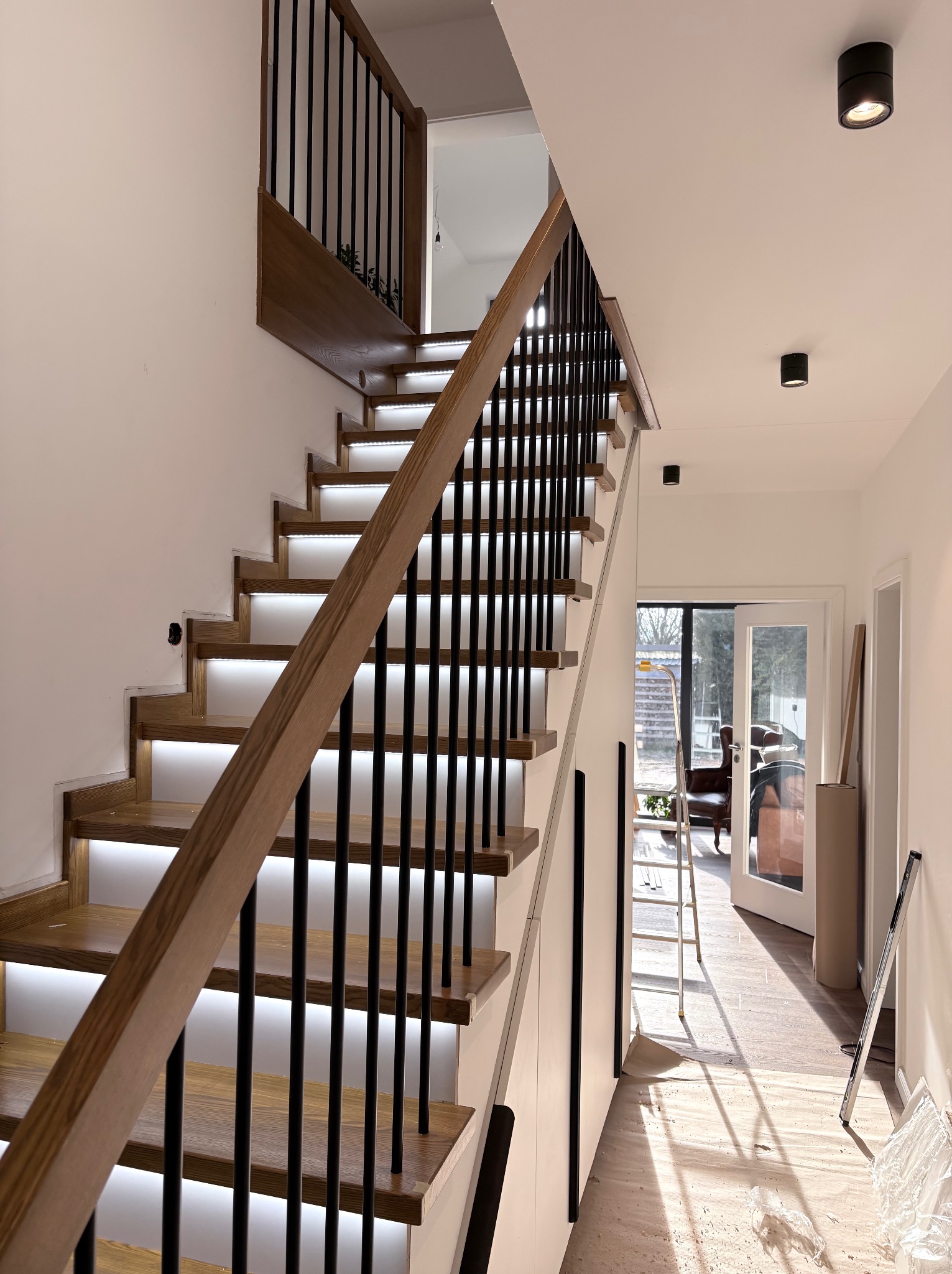 Nowoczesne schody drewniane z podświetleniem LED stopni i czarną balustradą w jasnym wnętrzu domu. Widoczny fragment salonu z fotelami i drabina malarska.