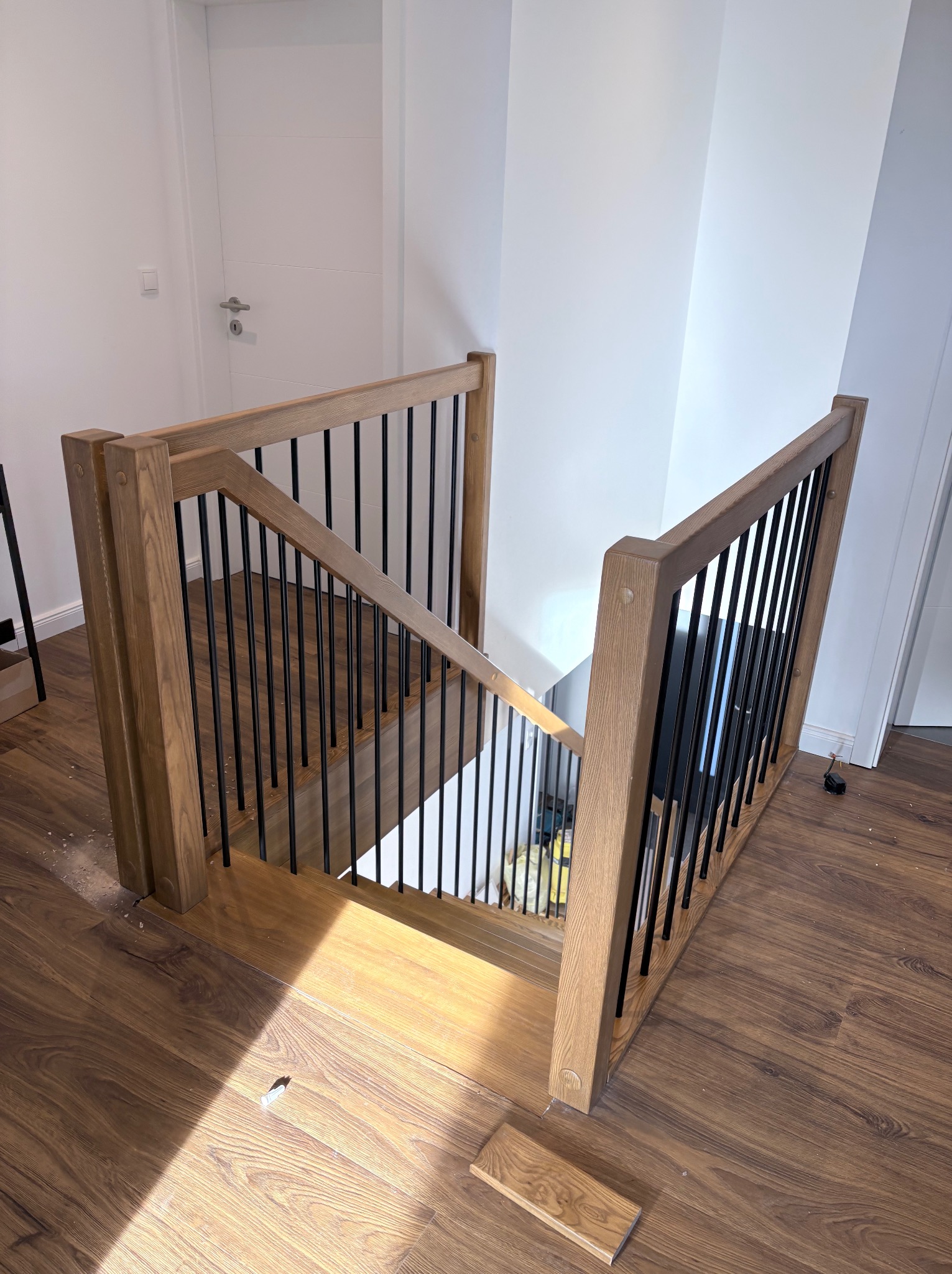 Drewniane schody z czarnymi, metalowymi balustradami, widok z góry, jasne wnętrze z drewnianą podłogą i białymi ścianami, fragment niedokończonej instalacji.