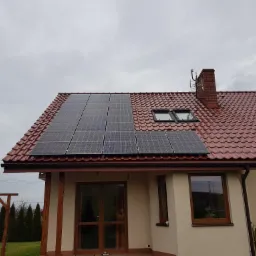 Panele słoneczne zamontowane na czerwonym dachu domu jednorodzinnego z kominem i oknami dachowymi, widok z poziomu ogrodu w pochmurny dzień.