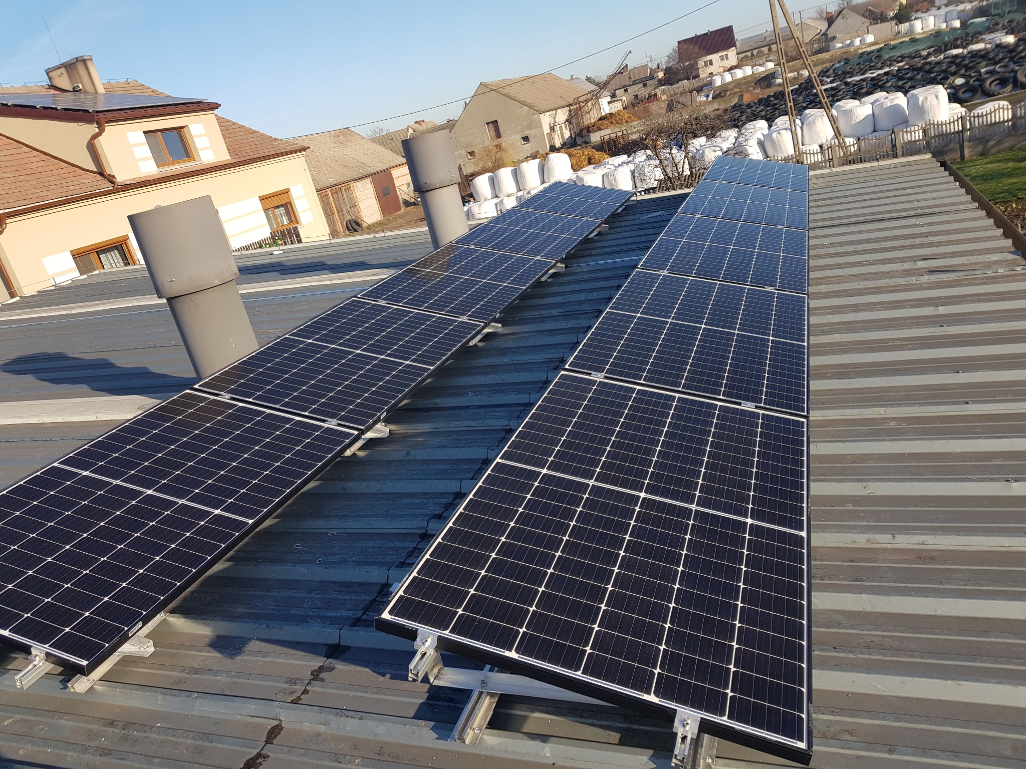 Rząd paneli słonecznych zamontowanych na szarym, blaszanym dachu budynku gospodarczego na wsi, w tle domy i stogi siana w białych workach.