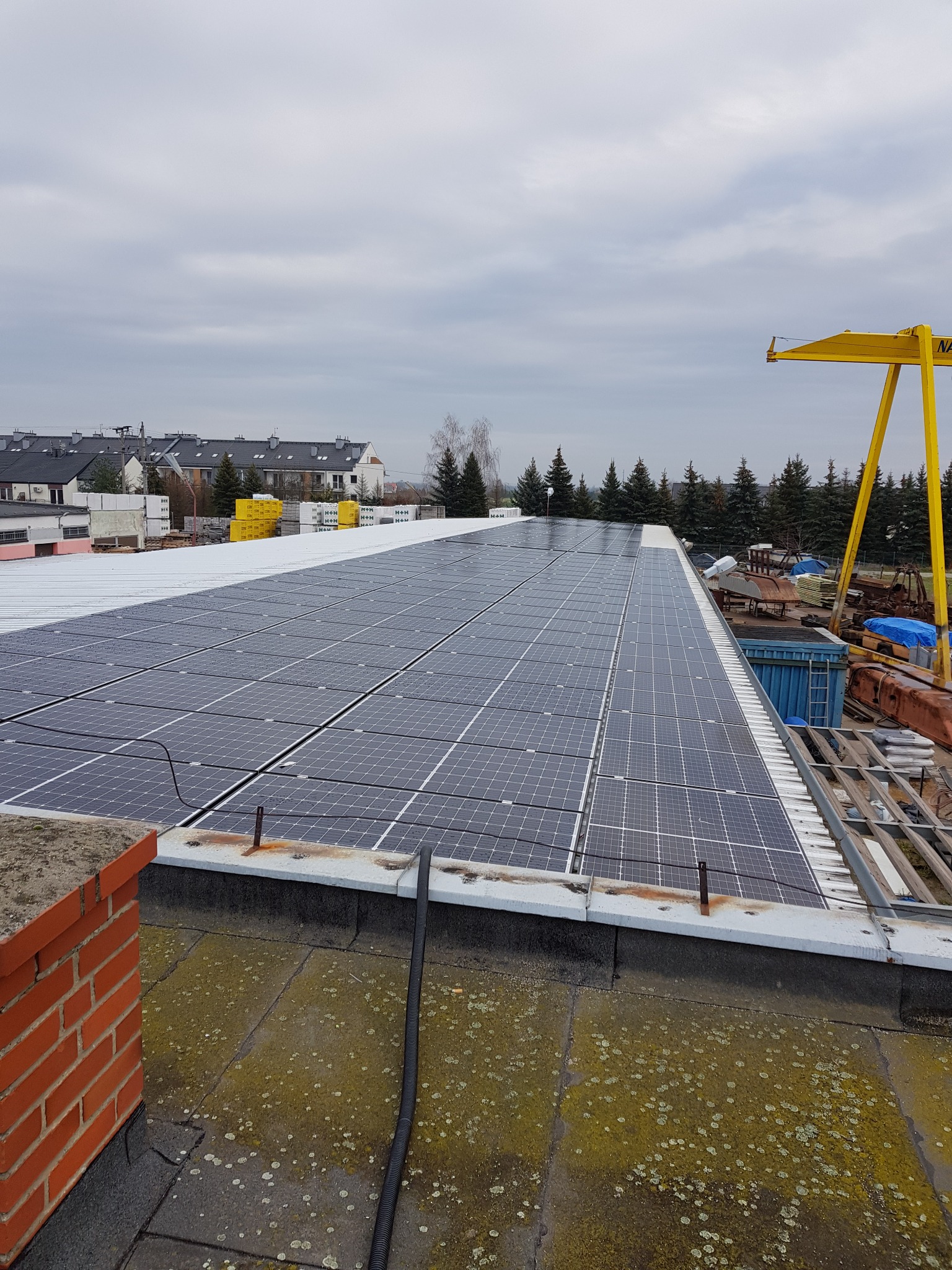 Rząd paneli słonecznych na płaskim dachu budynku przemysłowego, widoczne elementy konstrukcyjne dachu i żółty dźwig w tle, pochmurne niebo.