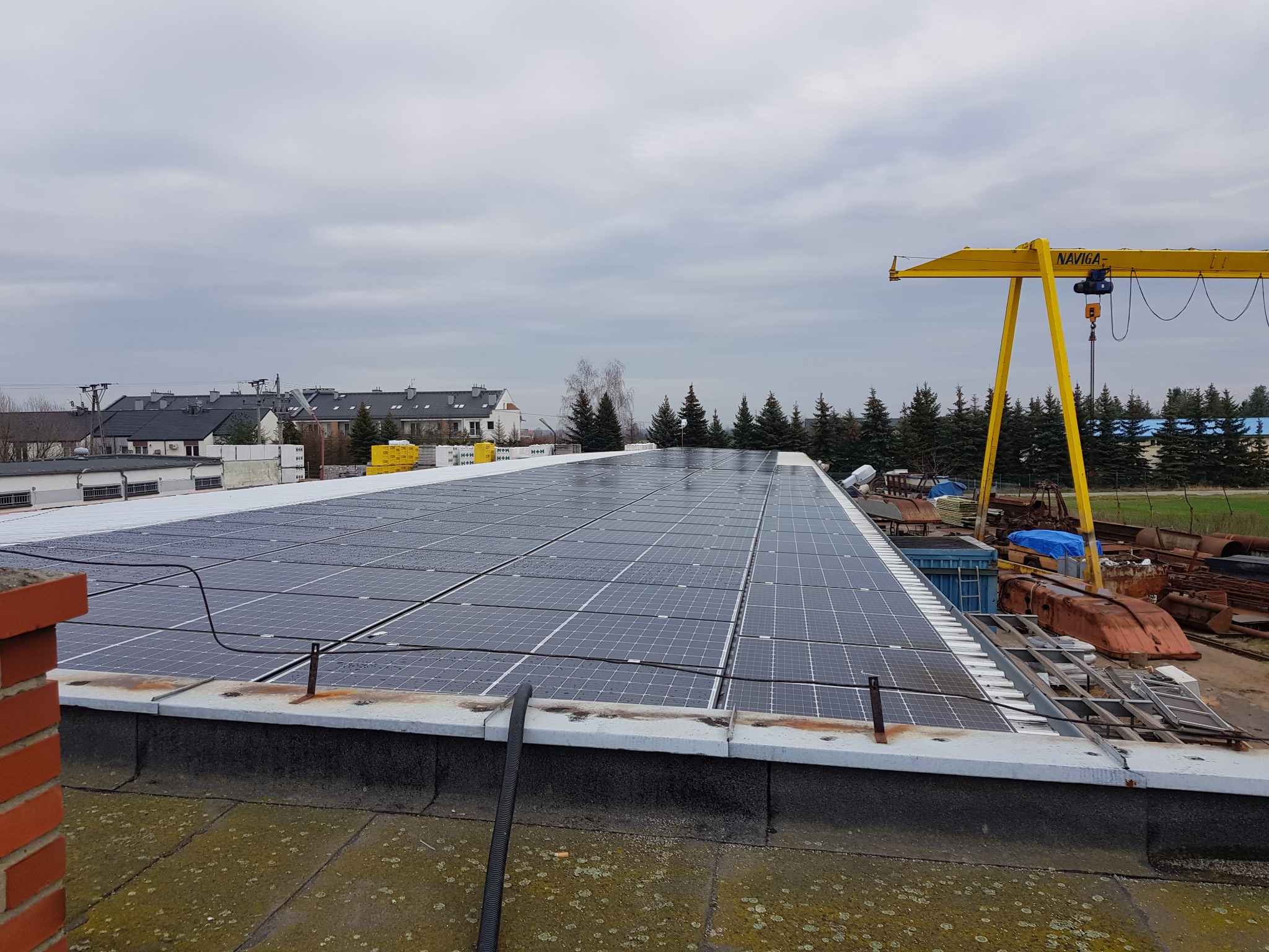 Rząd paneli słonecznych na płaskim dachu budynku przemysłowego, widoczny żółty dźwig bramowy i zabudowania w tle, pochmurne niebo.