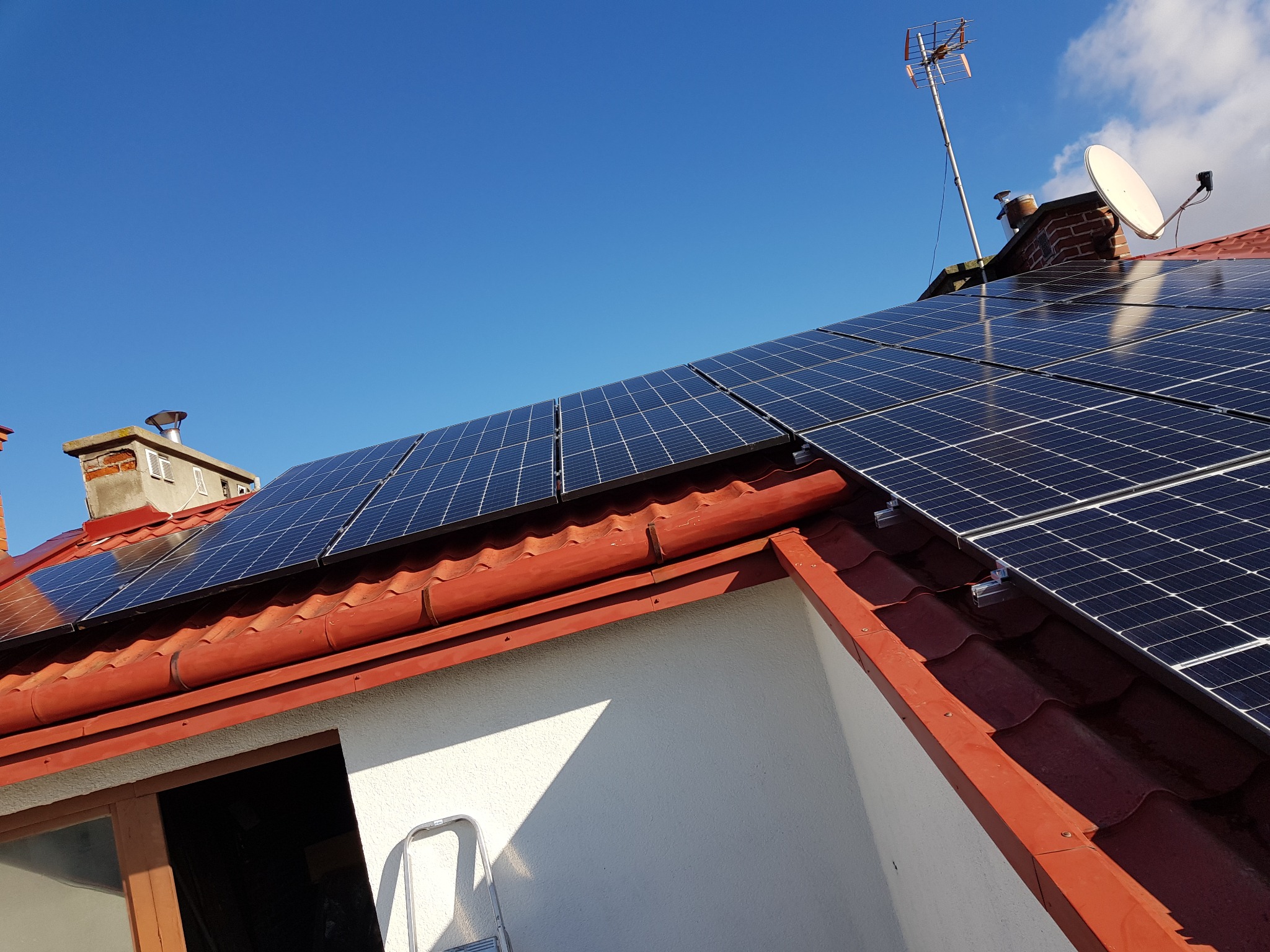 Instalacja fotowoltaiczna na dachu z czerwonej dachówki karpiówki, widoczna drabina oparta o ścianę budynku, antena telewizyjna na kominie w tle, słoneczny dzień.
