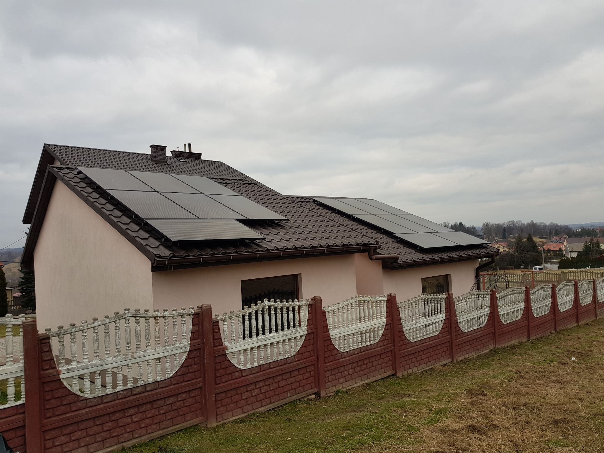 Dom jednorodzinny z panelami fotowoltaicznymi na dachu, widok z poziomu gruntu, ogrodzenie betonowe w pierwszym planie, pochmurne niebo.