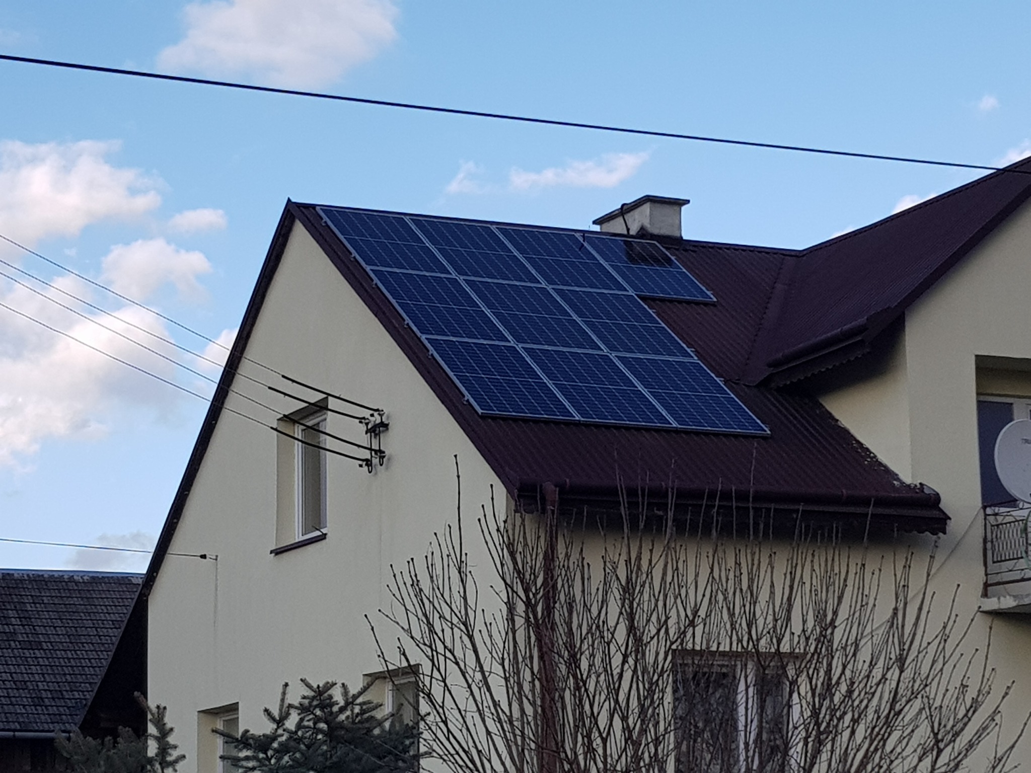 Panele słoneczne na dwuspadowym dachu domu jednorodzinnego z brązową blachodachówką, widoczne kable elektryczne i antena satelitarna na balkonie sąsiedniego budynku.