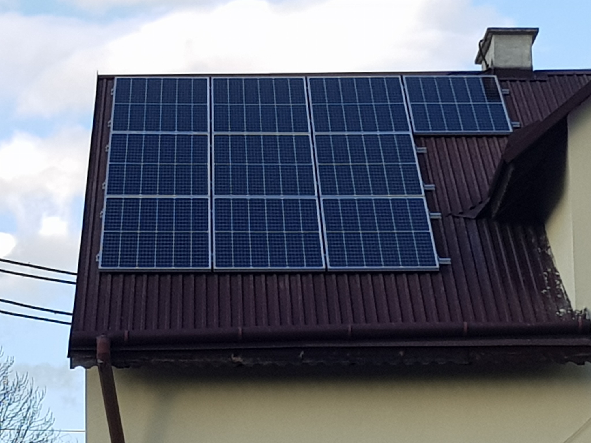 Panele słoneczne zamontowane na brązowym dachu domu jednorodzinnego, widoczne rury spustowe i komin, częściowo pochmurne niebo w tle.