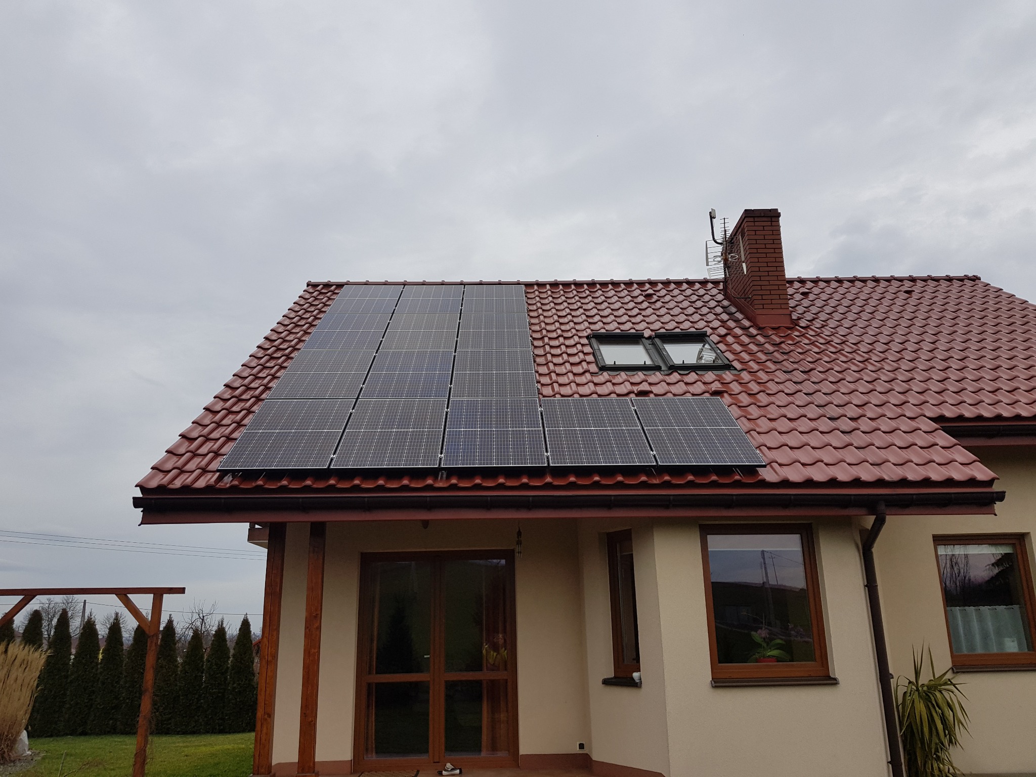 Panele słoneczne zamontowane na czerwonym dachu domu jednorodzinnego z kominem i oknami dachowymi, widok z poziomu ogrodu w pochmurny dzień.
