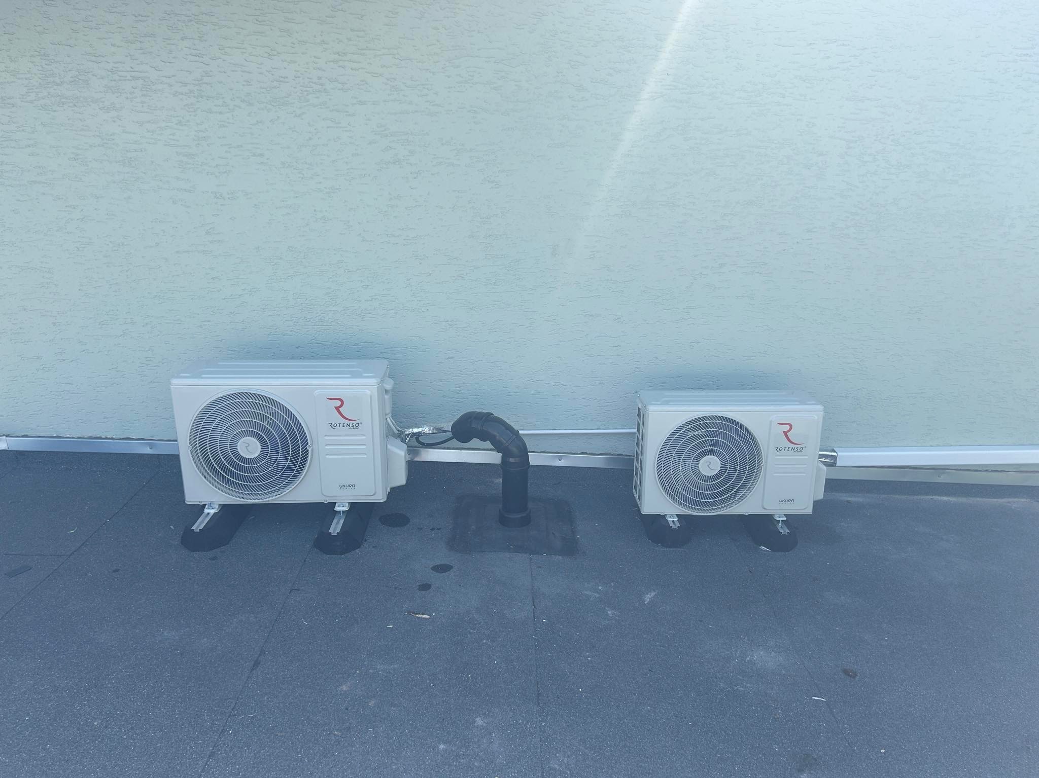 Dwa białe klimatyzatory marki Rotenso Ukura zamontowane na płaskim dachu, z widocznym czarnym przewodem odprowadzającym skropliny.