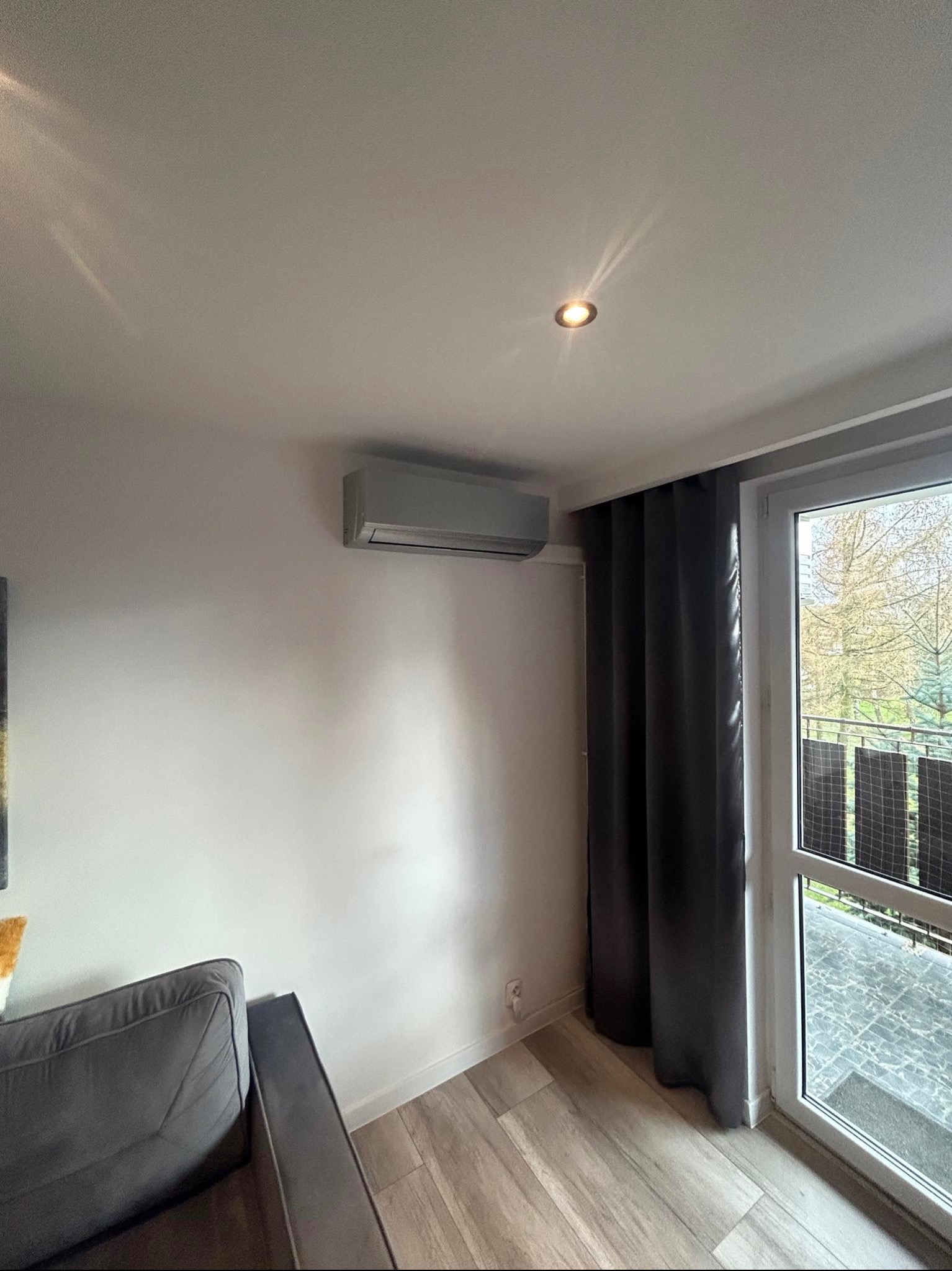 Szary klimatyzator ścienny w nowoczesnym salonie z drewnianą podłogą, zasłoniętym oknem balkonowym i widokiem na zewnątrz.