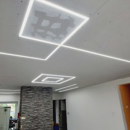 PRO-BUD COMPLEX - Sufit z płyt gipsowych z kwadratowymi lampami LED, częściowo wyszpachlowany, widok z dołu, stół z narzędziami w tle.