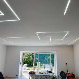 PRO-BUD COMPLEX - Nowoczesne oświetlenie sufitowe LED w kształcie geometrycznym, widok na taras przez otwarte drzwi balkonowe, pomieszczenie w trakcie wykończenia.