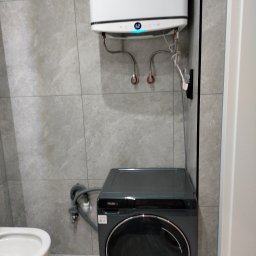 PRO-BUD COMPLEX - Kompaktowa łazienka z szarą pralką Haier i białym bojlerem Ariston na tle płytek imitujących kamień. Widoczne przyłącza wodne i odpływ. Minimalistyczny design.