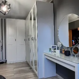 Biała szafa z lustrami i toaletka z okrągłym lustrem w nowoczesnym pokoju z panelami podłogowymi w odcieniach szarości i beżu.