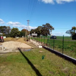 Fragment budowy ogrodzenia z siatki, widoczne słupki przygotowane do montażu, pryzma piasku i traktor w tle, słoneczny dzień.