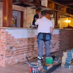 Montaż dekoracyjnej ściany z cegły w lokalu, widoczny fragment baru z dwoma osobami, narzędzia i materiały budowlane, poziomica laserowa wyznacza linię.