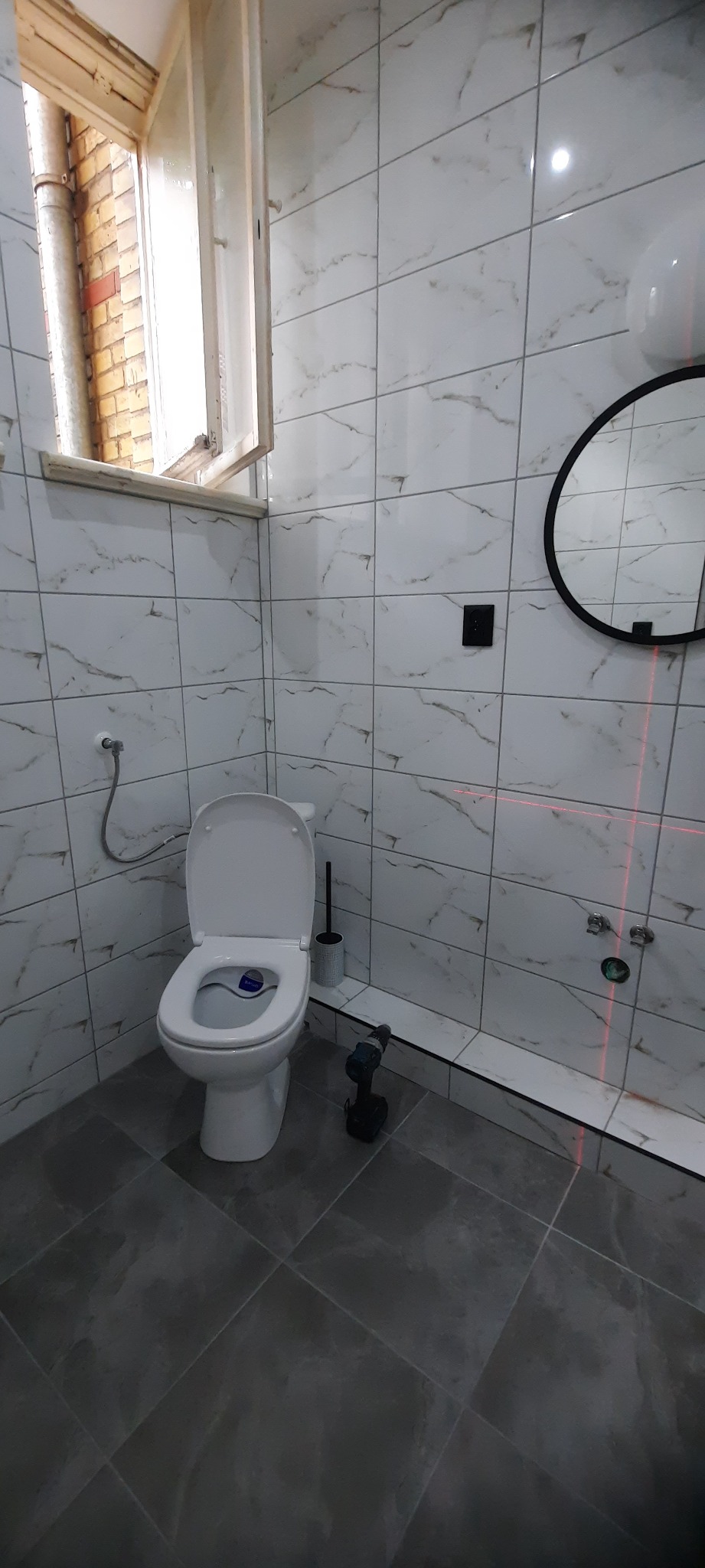 Nowoczesna łazienka z białymi marmurkowymi płytkami ściennymi, ciemnoszarą podłogą, toaletą z otwartą klapą, okrągłym lustrem i poziomnicą laserową, w trakcie prac wykończeniowych, widoczna...