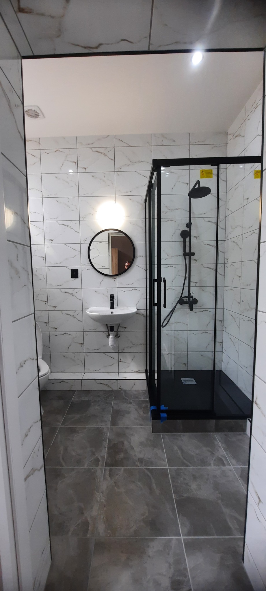 Nowoczesna łazienka z czarną kabiną prysznicową, okrągłym lustrem z podświetleniem i umywalką na tle marmurowych płytek.