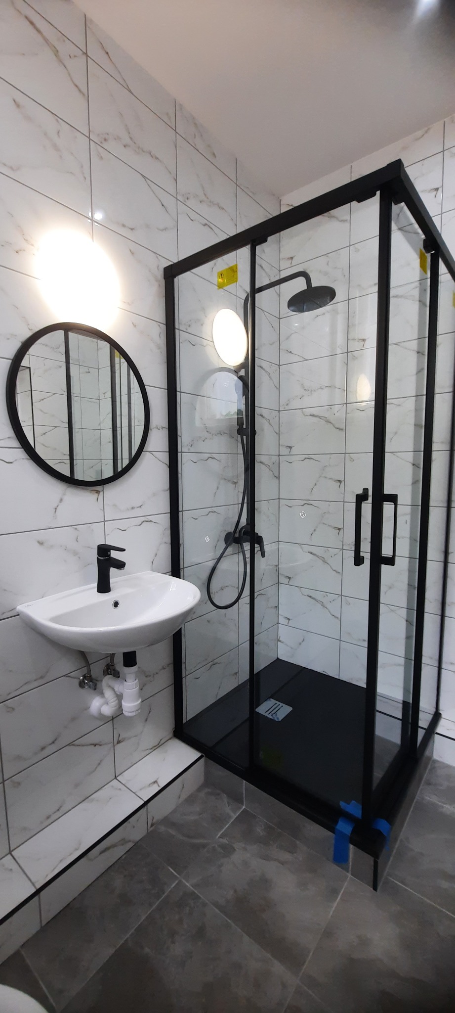 Nowoczesna łazienka z czarną kabiną prysznicową typu walk-in, okrągłym lustrem nad białą umywalką z czarną baterią, białe płytki imitujące marmur na ścianach i szare płytki na podłodze.