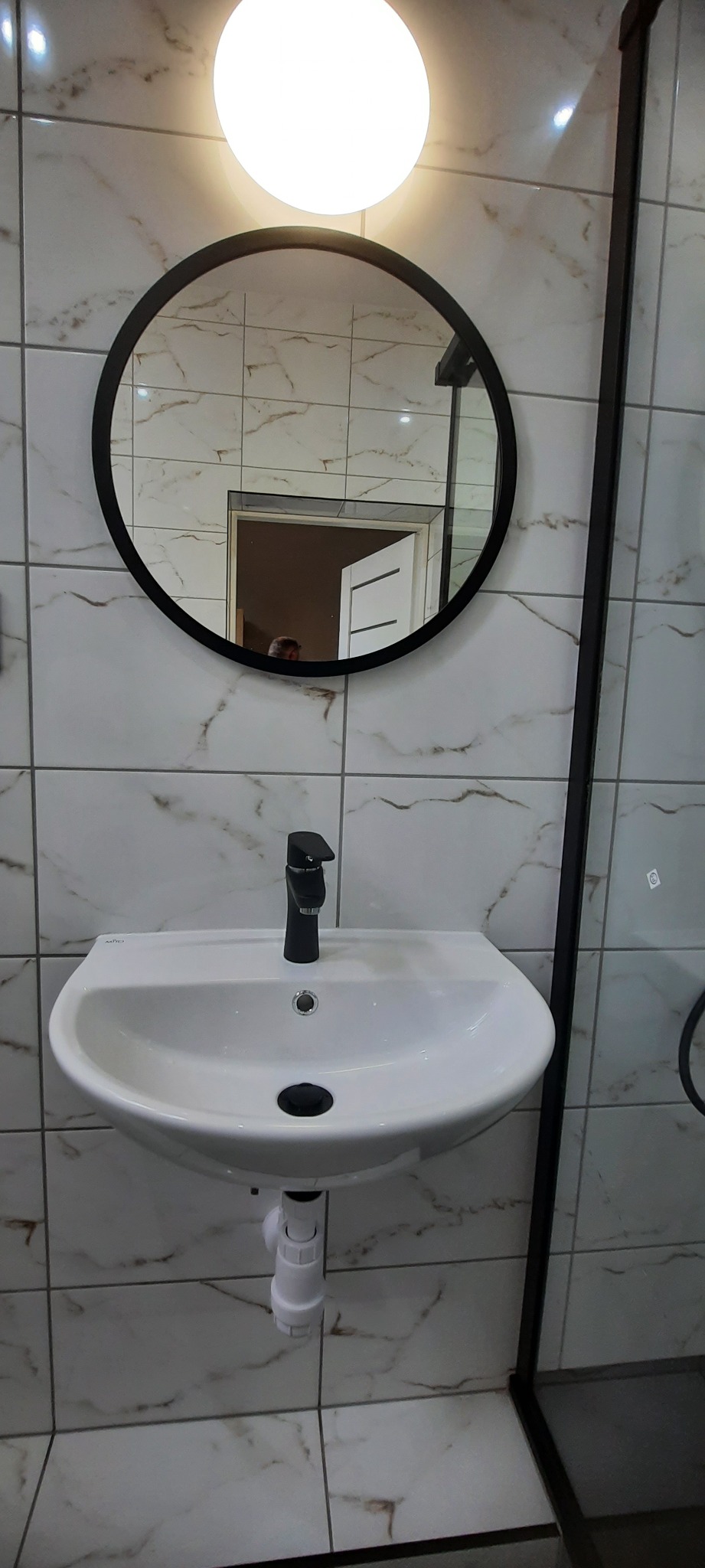 Umywalka nablatowa z czarną baterią, okrągłe lustro z czarną ramą i oświetlenie sufitowe w łazience wykończonej marmurkowymi płytkami.