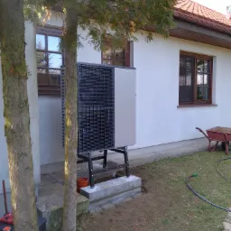 Zewnętrzna jednostka pompy ciepła zamontowana na betonowym fundamencie obok drzewa, z taczką ziemi i wężem ogrodowym w tle.