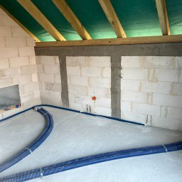 W.R. Silver System - Wnętrze budynku w trakcie budowy z widocznymi niebieskimi rurami instalacji grzewczej lub wodnej ułożonymi na podłodze, ściany z bloczków betonowych i drewniany szkielet dachu z zieloną membraną.