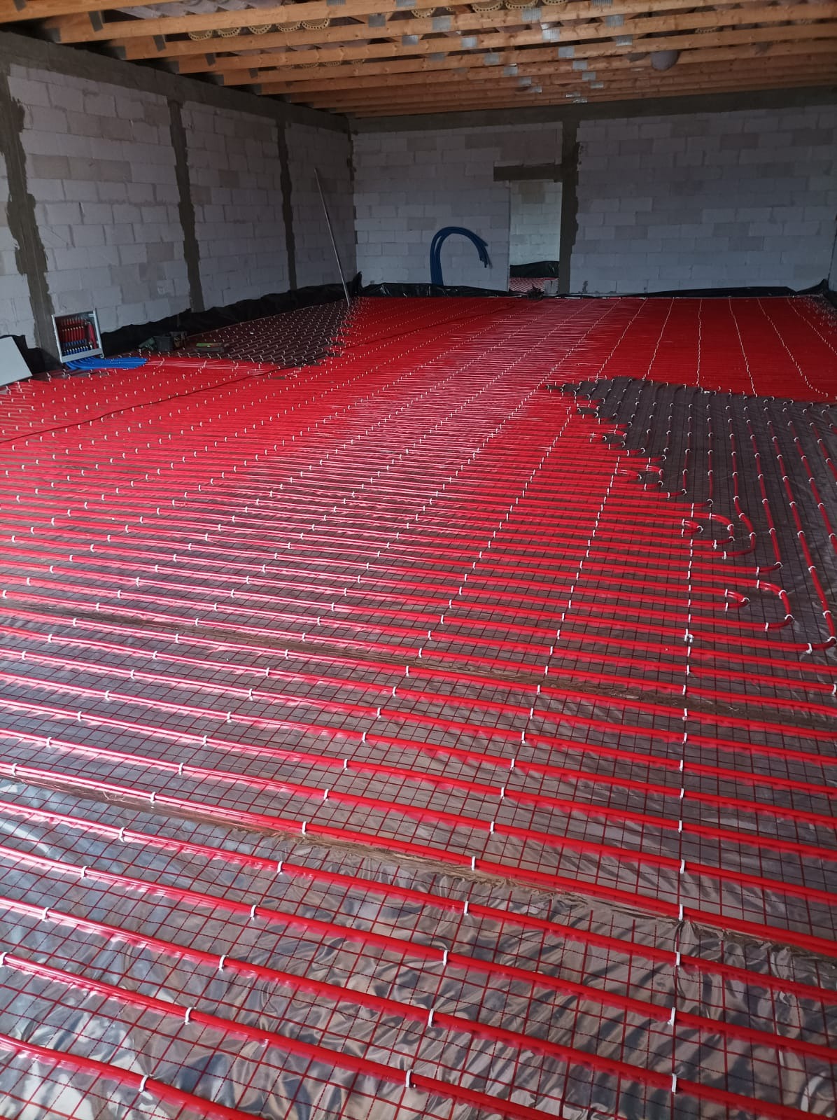 Czerwone rurki ogrzewania podłogowego rozłożone na folii izolacyjnej w surowym pomieszczeniu z betonowymi ścianami i drewnianym stropem.