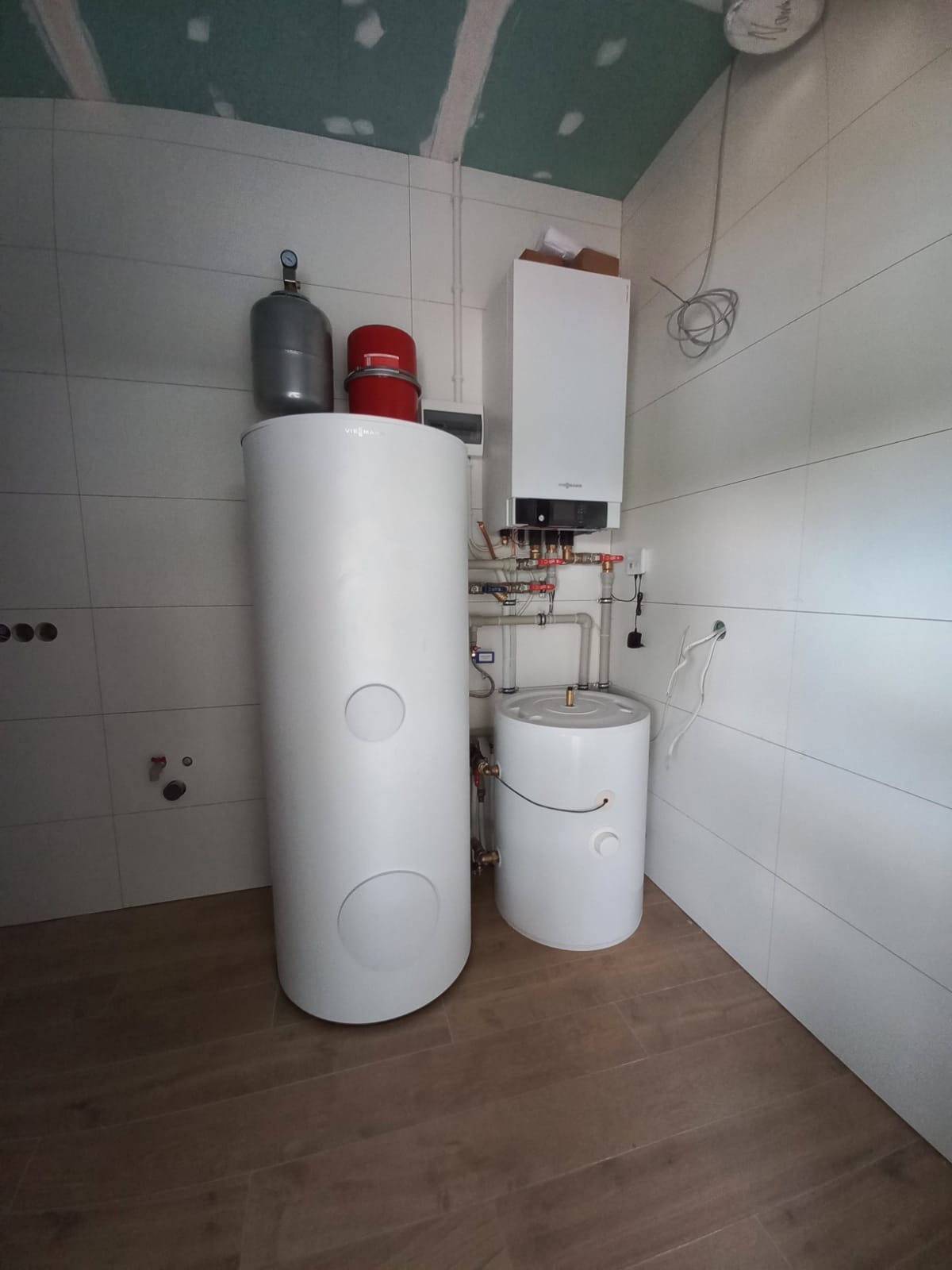 Biały zasobnik ciepłej wody użytkowej, szary zbiornik wyrównawczy, czerwony naczynie przeponowe i wiszący kocioł gazowy Viessmann zamontowane w pomieszczeniu z wykończonymi płytkami ścianami...