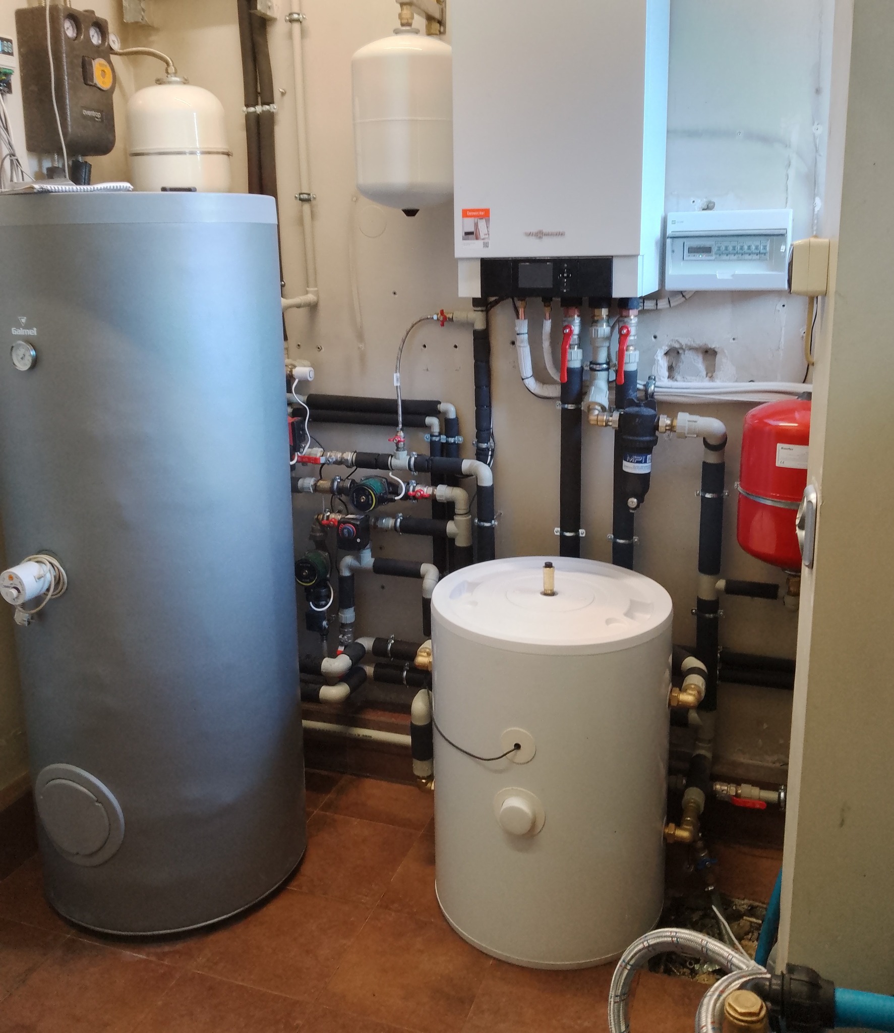 Instalacja kotła gazowego Viessmann z dwoma zasobnikami wody, widoczne liczne rury i zawory, całość w pomieszczeniu z brązowymi płytkami podłogowymi.