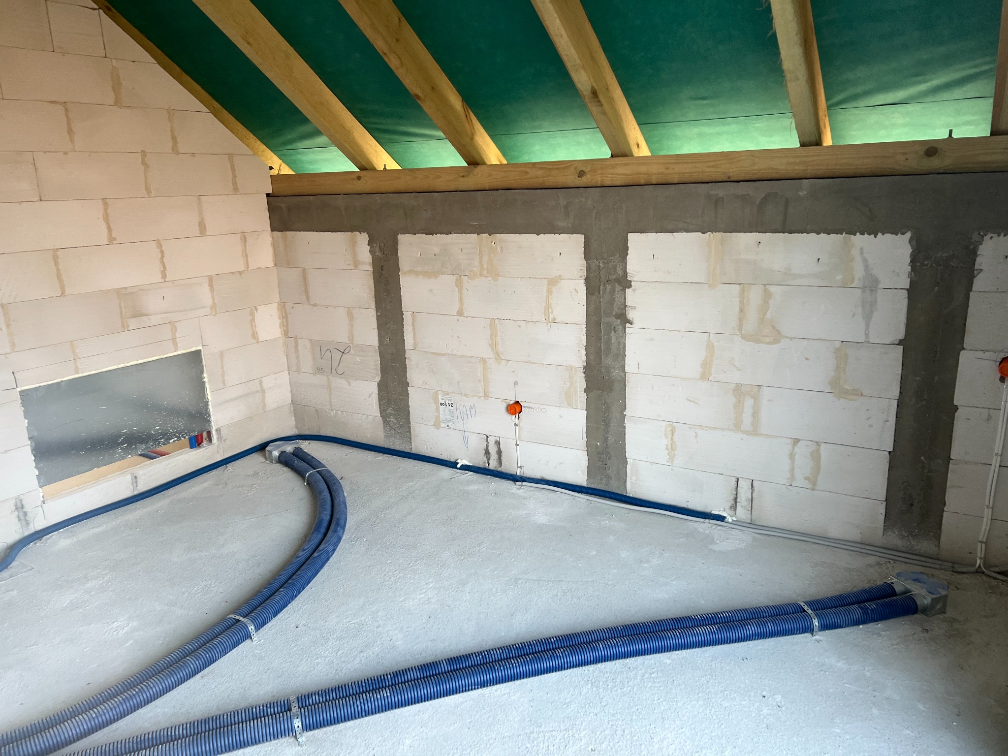Wnętrze budynku w trakcie budowy z widocznymi niebieskimi rurami instalacji grzewczej lub wodnej ułożonymi na podłodze, ściany z bloczków betonowych i drewniany szkielet dachu z zieloną membraną.