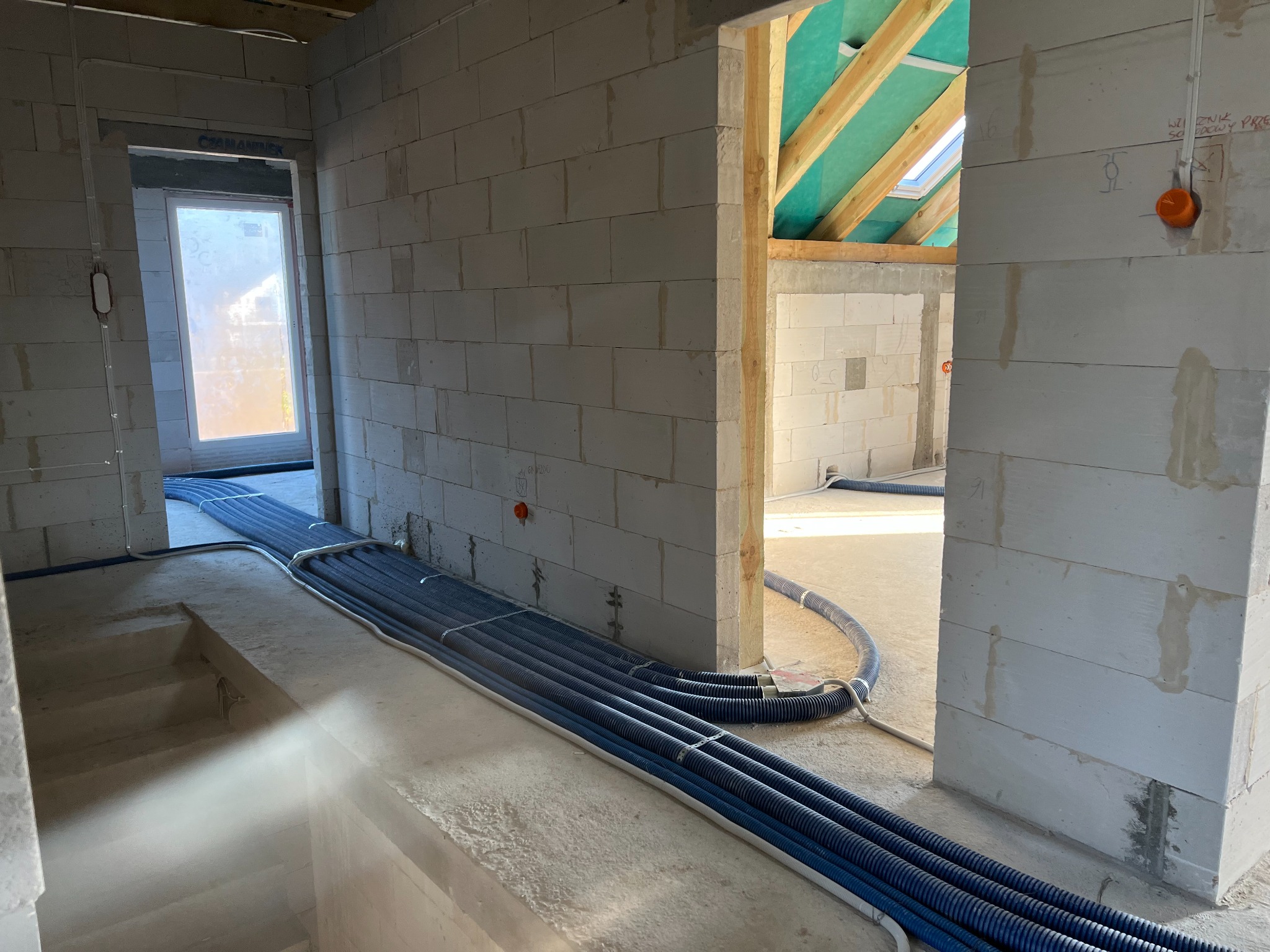 Wnętrze w surowym stanie z widocznymi niebieskimi rurami instalacyjnymi ułożonymi wzdłuż ściany i podłogi, prowadzącymi do otworu drzwiowego; widoczne ściany z betonu komórkowego i drewniana...