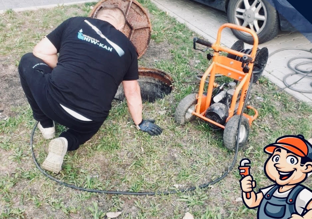 Hydraulik w czarnej koszulce z logo firmy, klęczy przy otwartym włazie kanalizacyjnym. Obok stoi pomarańczowa maszyna do udrażniania rur.