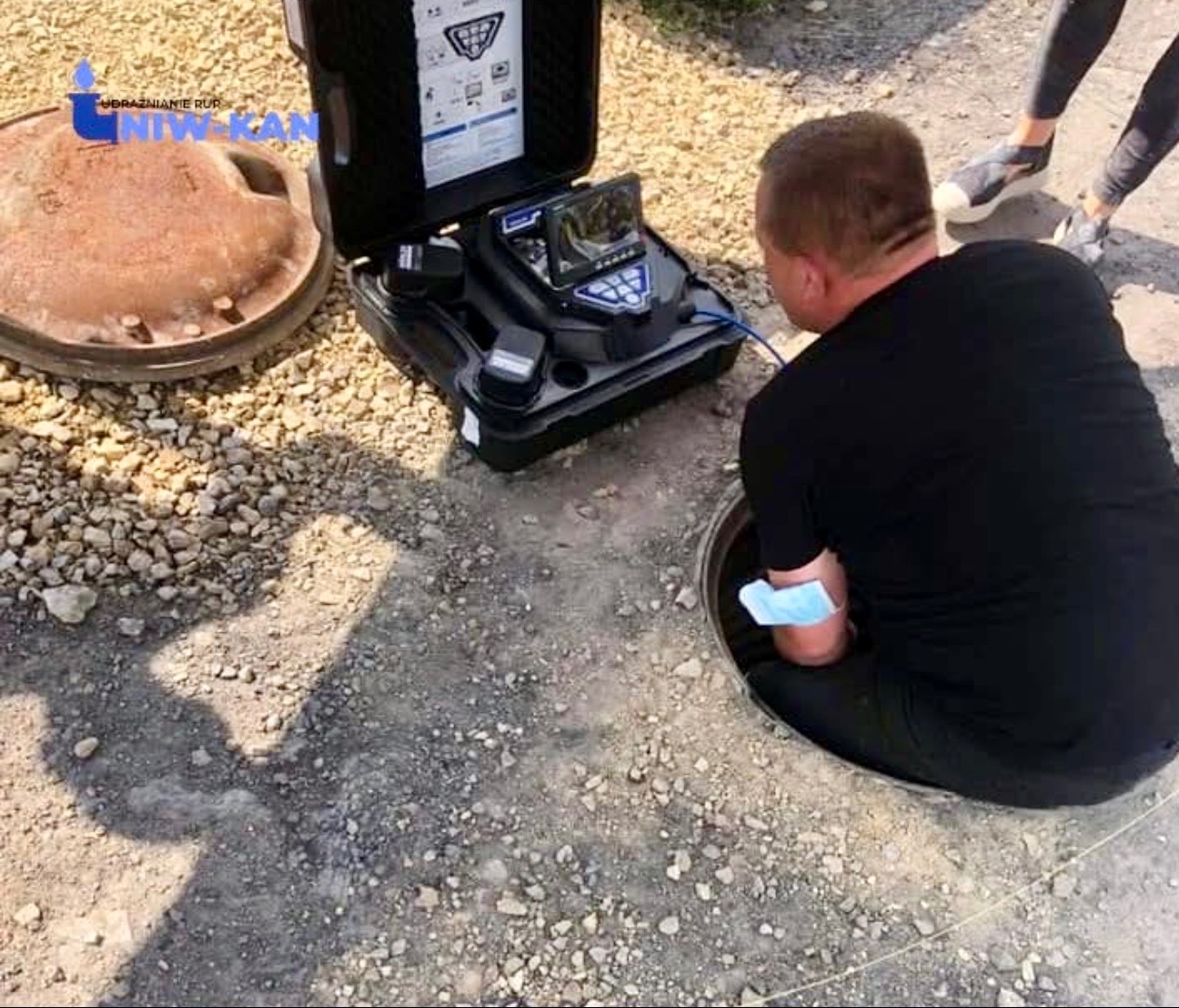Hydraulik z kamerą inspekcyjną zagląda do studzienki kanalizacyjnej. Obok leży pokrywa i stoi otwarty sprzęt diagnostyczny. Widoczne nogi drugiej osoby.