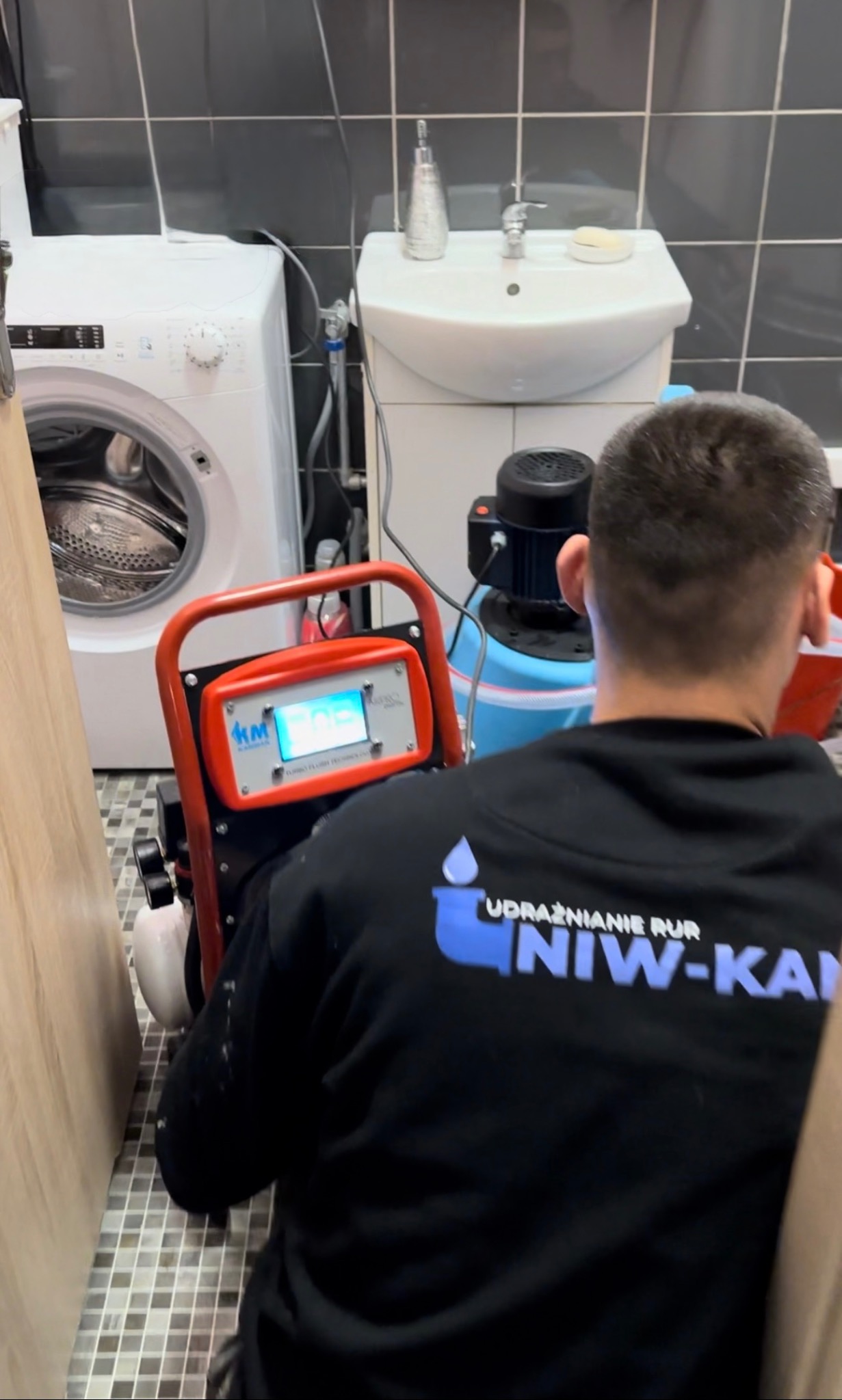 Hydraulik z urządzeniem do udrażniania rur w łazience z pralką i umywalką. Na bluzie widoczne logo firmy 'Udrażnianie Rur NIW-KAN'.