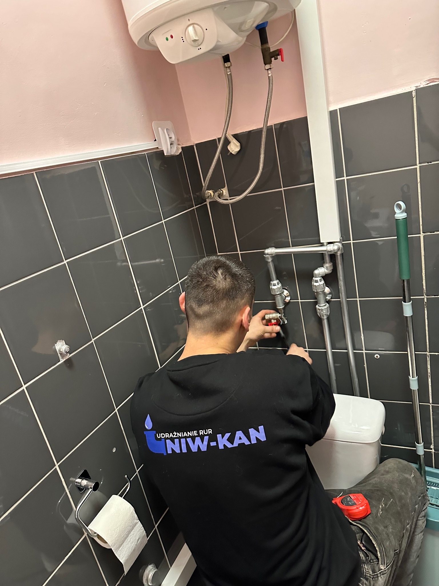 Hydraulik w trakcie pracy nad instalacją wodną w łazience z szarymi płytkami. Widoczny bojler, rury i narzędzia. Na bluzie logo firmy.