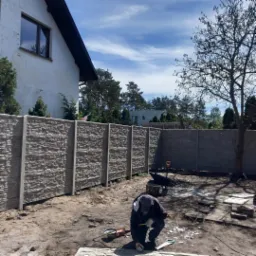 Montaż prefabrykowanego ogrodzenia betonowego z teksturą kamienia, pracownik docina element na kolanach, widoczne narzędzia i taczka na piaszczystym podłożu, w tle dom jednorodzinny i drzewa.