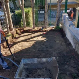 Przygotowany teren pod przyszłą nawierzchnię, widoczna wytyczona nić, częściowo ułożone obrzeża, taczka z ziemią i pracownik z łopatą, w tle budowa murku z bloczków.