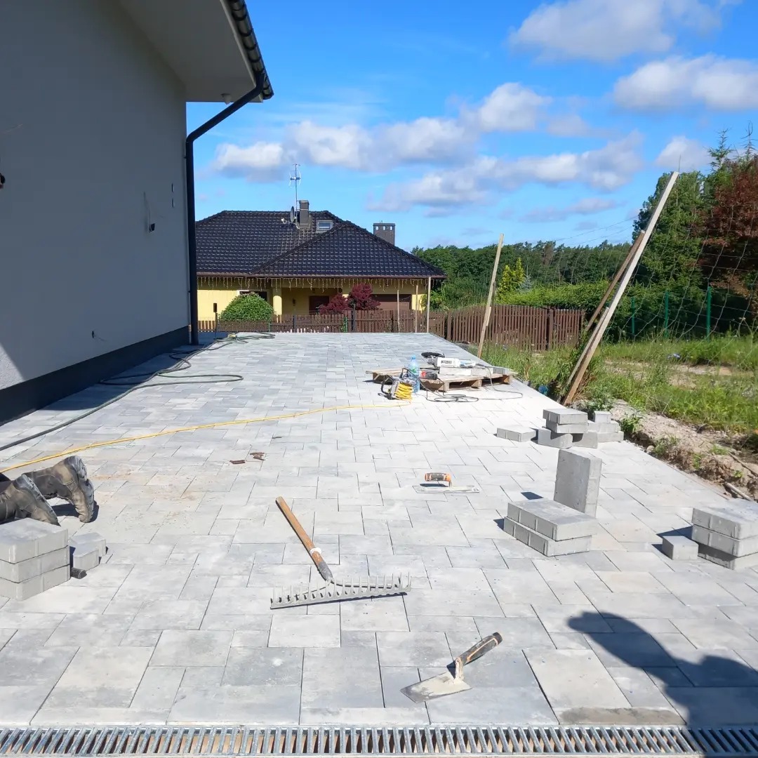Szara kostka brukowa ułożona na podjeździe domu jednorodzinnego, widoczne narzędzia brukarskie: grabie, kielnia, poziomica oraz paleta z materiałami budowlanymi w tle.