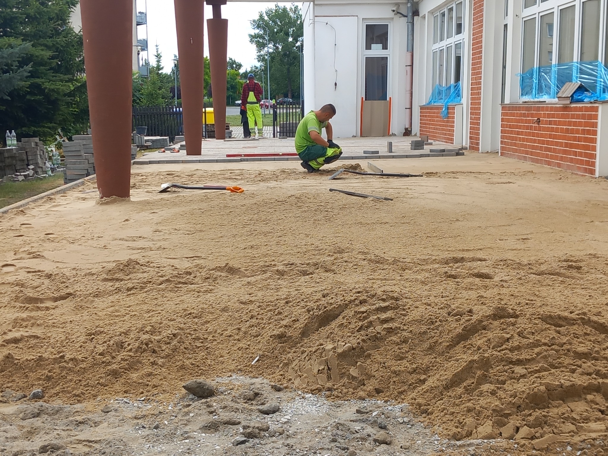 Pracownik w zielonym ubraniu klęczy na piasku, wyrównując podłoże pod kostkę brukową, widoczne narzędzia, częściowo ułożona kostka, budynek w tle z zabezpieczonymi oknami.